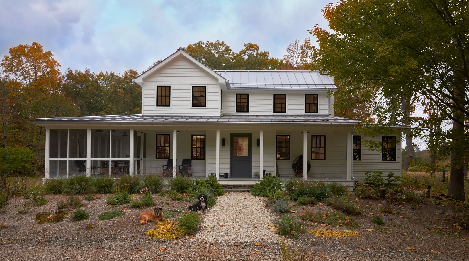 white farmhouse with a wraparound porch and metal roof, set among autumn trees with a gravel path to the door