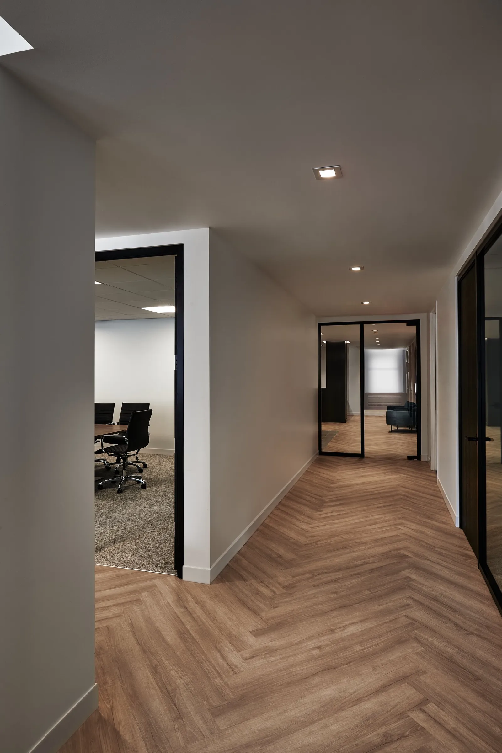 herringbone wood floor hallway with glass-walled meeting room on the left and glass doors ahead