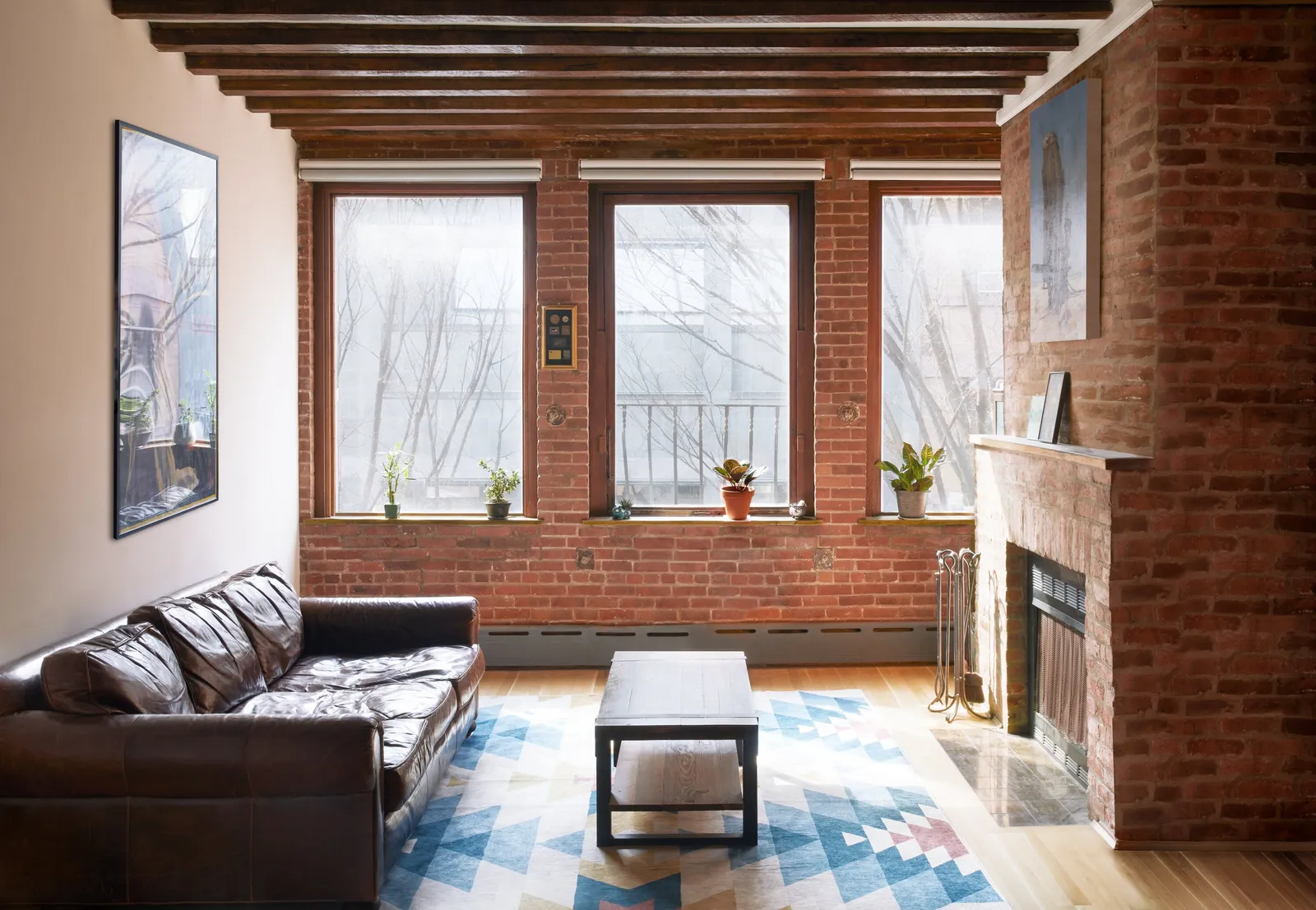 living room with exposed brick walls, large windows, a brown leather sofa, a blue patterned rug and a brick fireplace