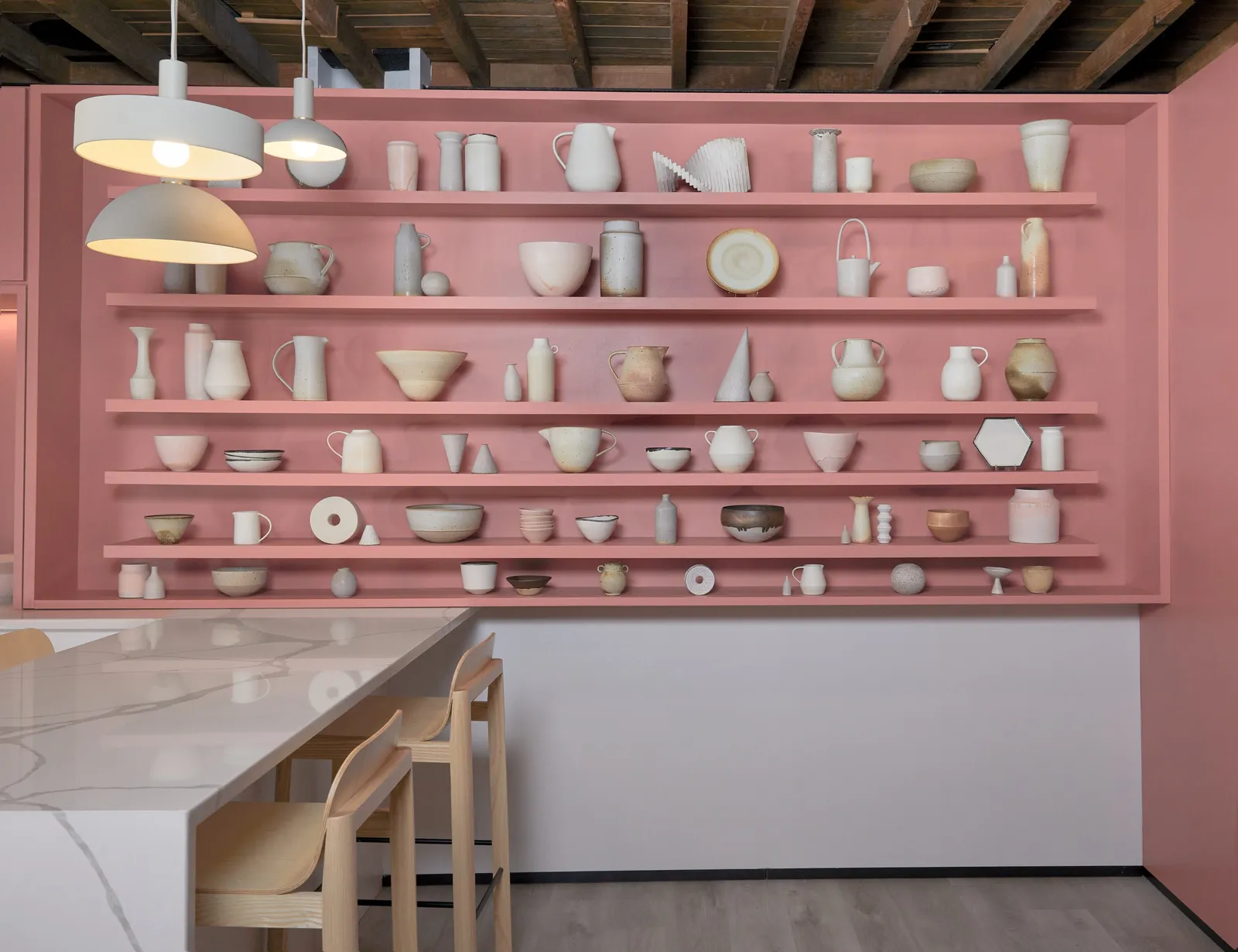 wall of pink open shelves filled with white and beige ceramics, pendant lamps and a marble table with wood stools