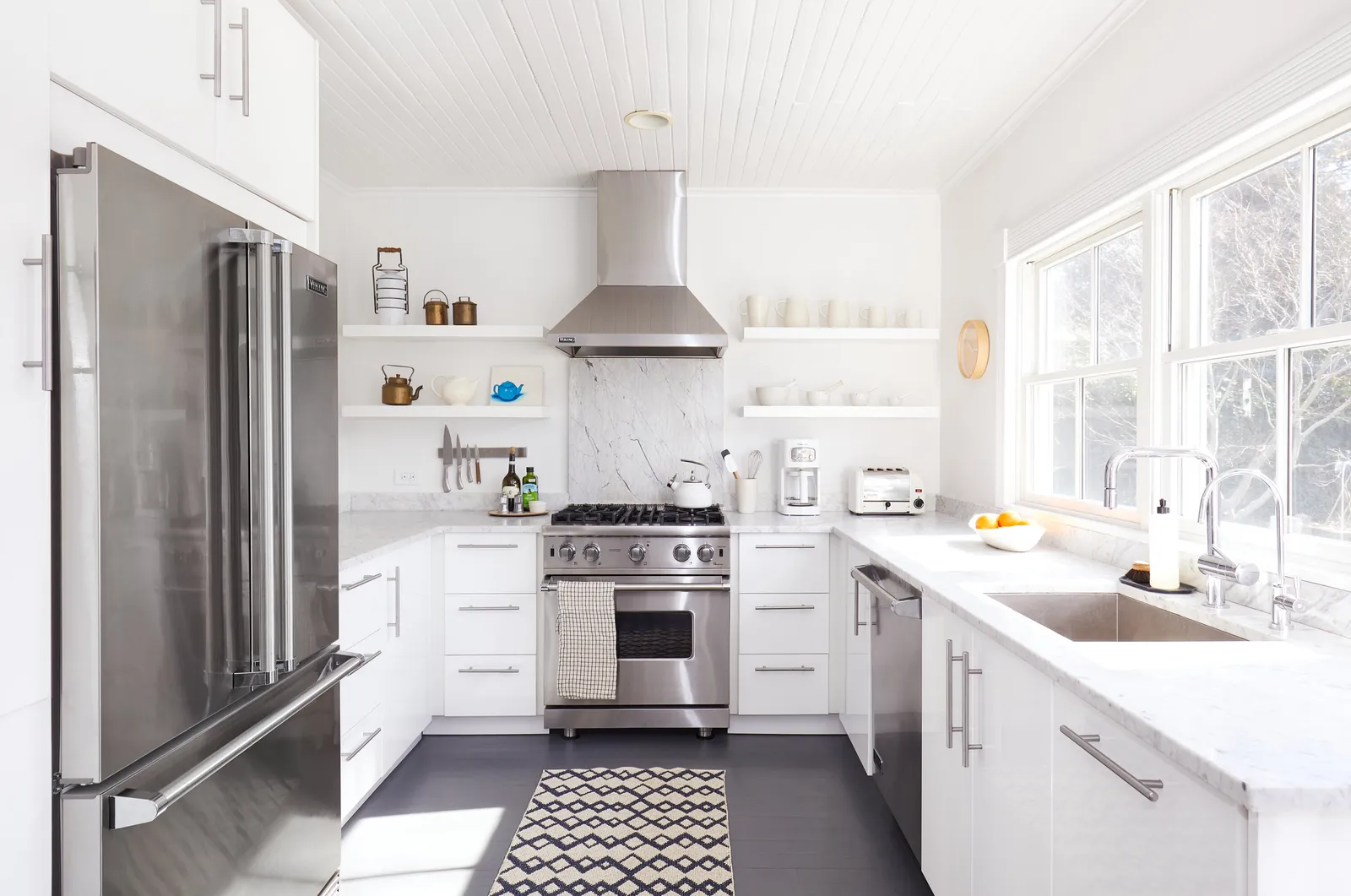 white kitchen with stainless appliances, open shelves, a marble counter and a graphic rug on a dark floor