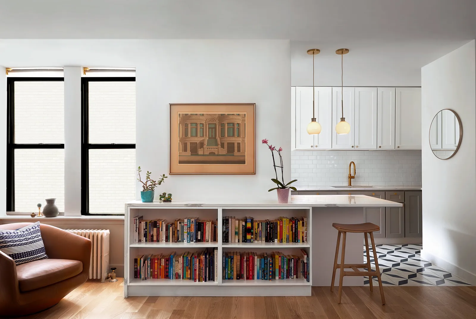 living and kitchen area with a low bookshelf peninsula, a leather armchair, framed print and brass pendant lights