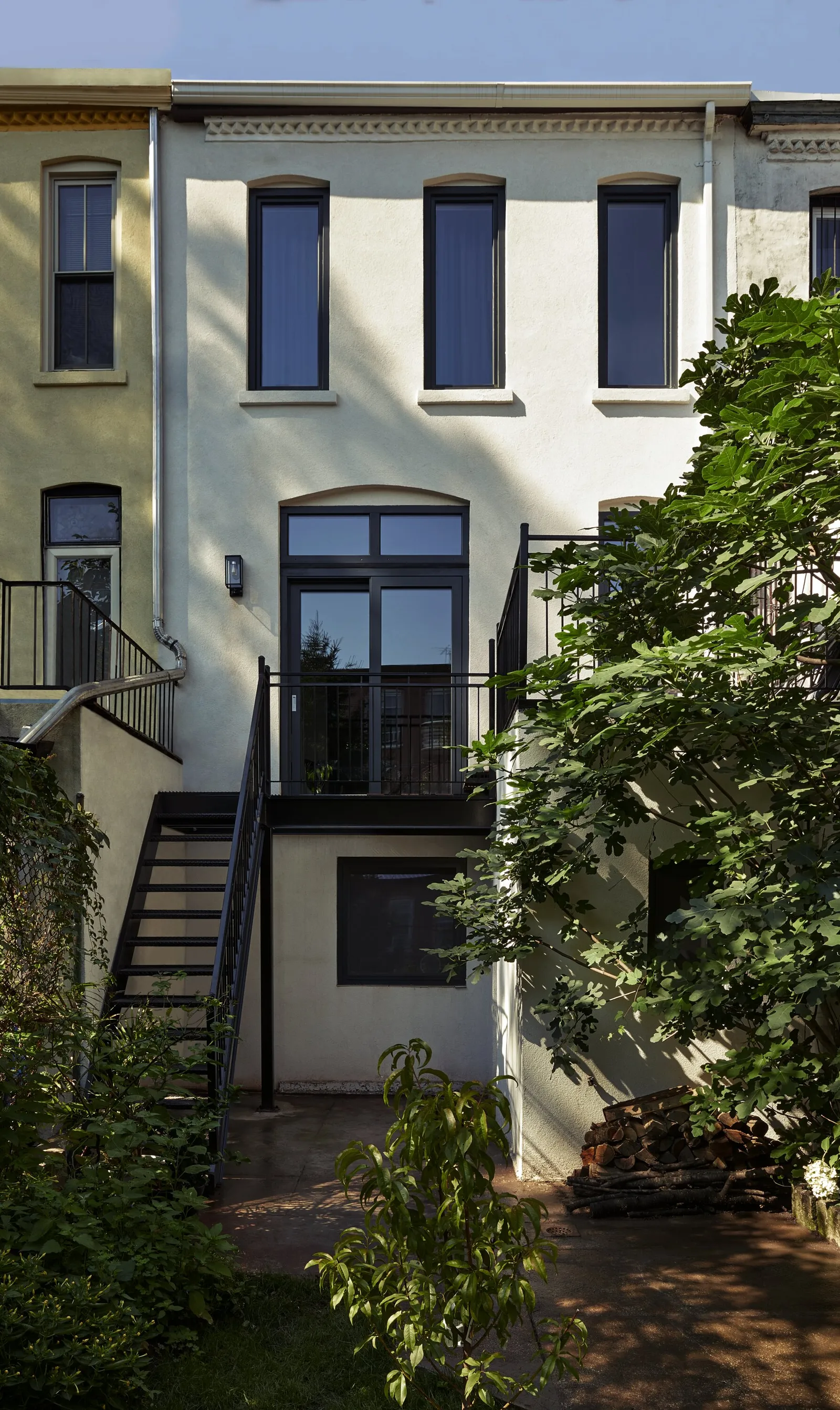 rear facade of a white townhouse with tall dark windows, an iron stair and a leafy tree in the garden