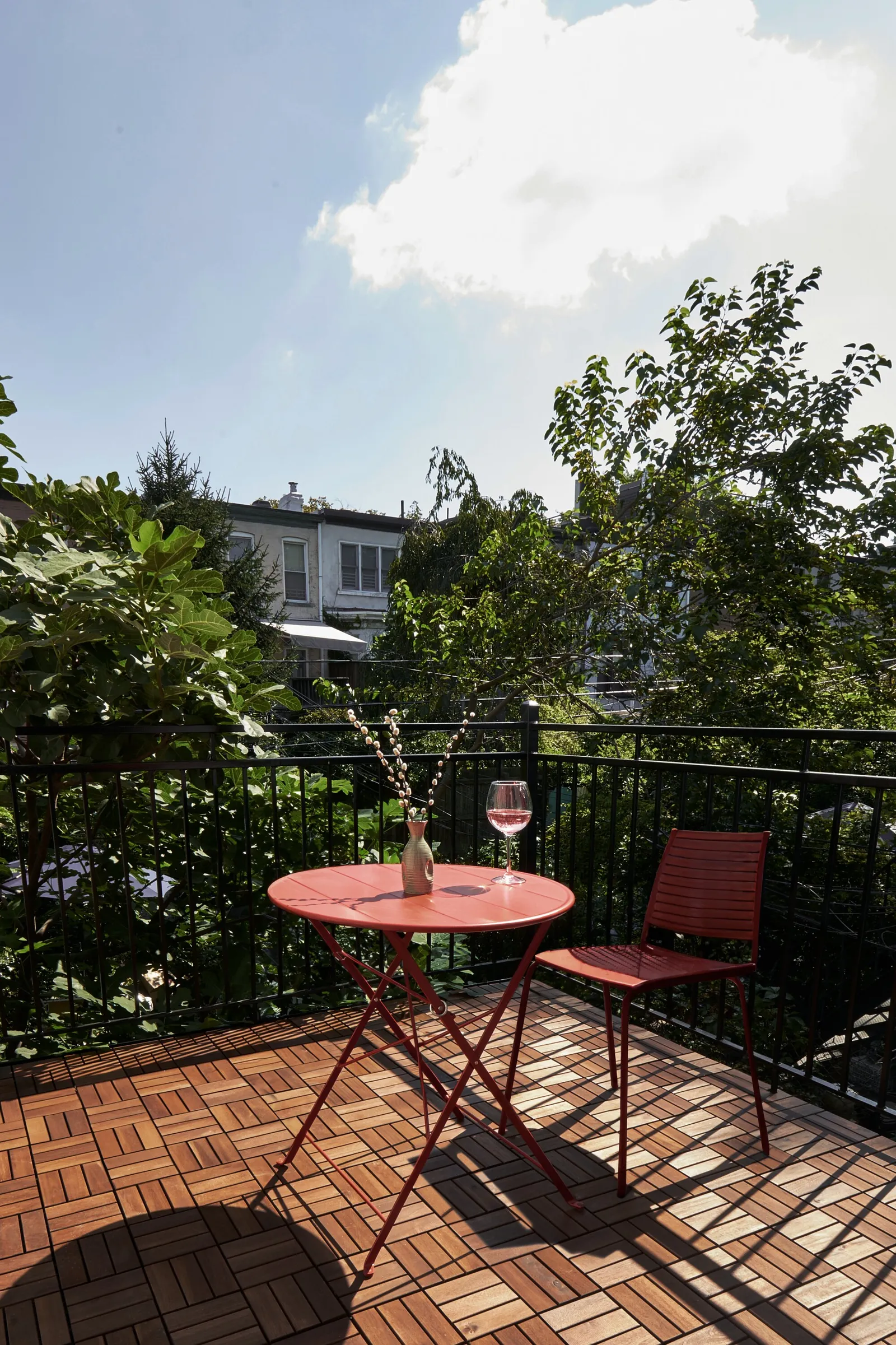 sunny wood-tiled deck with a small red bistro table and chair, a wine glass and a leafy view behind the railing