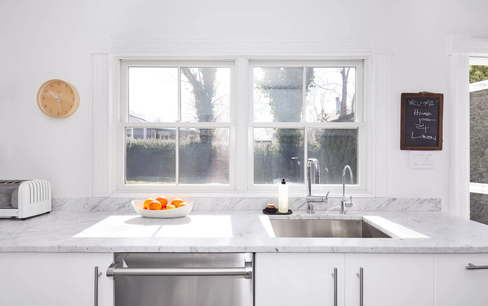 kitchen sink under a double window, with a bowl of oranges, a wooden wall clock and a small chalkboard