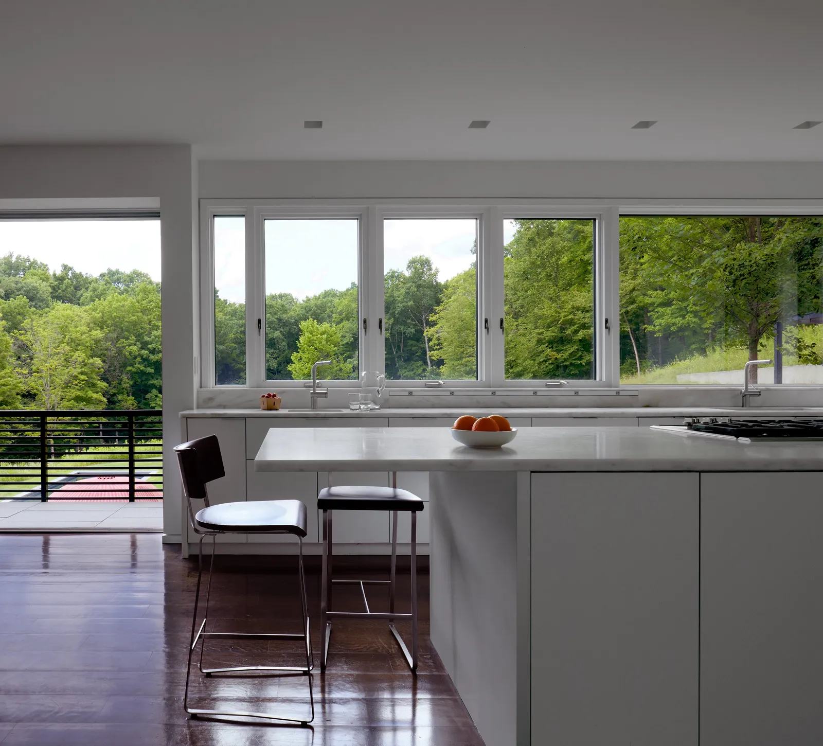 kitchen island with two leather stools and a bowl of oranges before a row of windows onto green trees