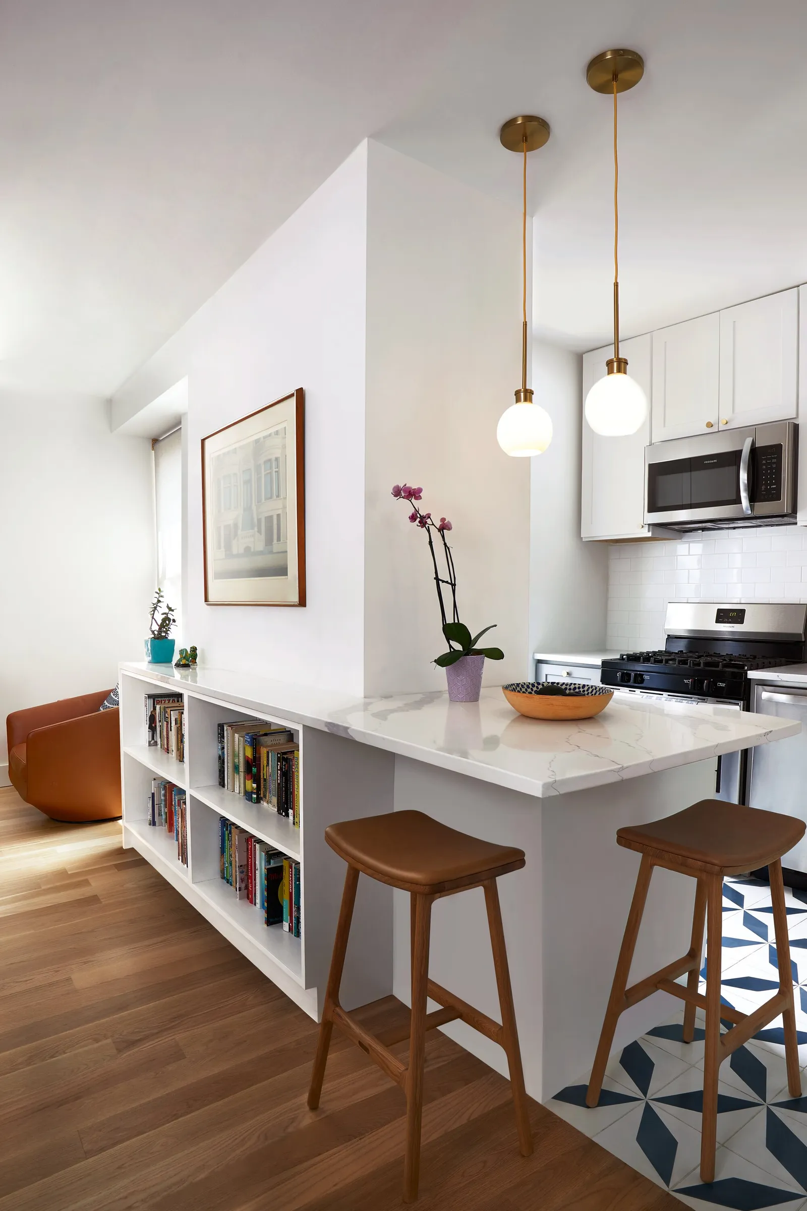 kitchen counter extending from a white bookcase, with two wooden stools, brass pendants and patterned floor tile