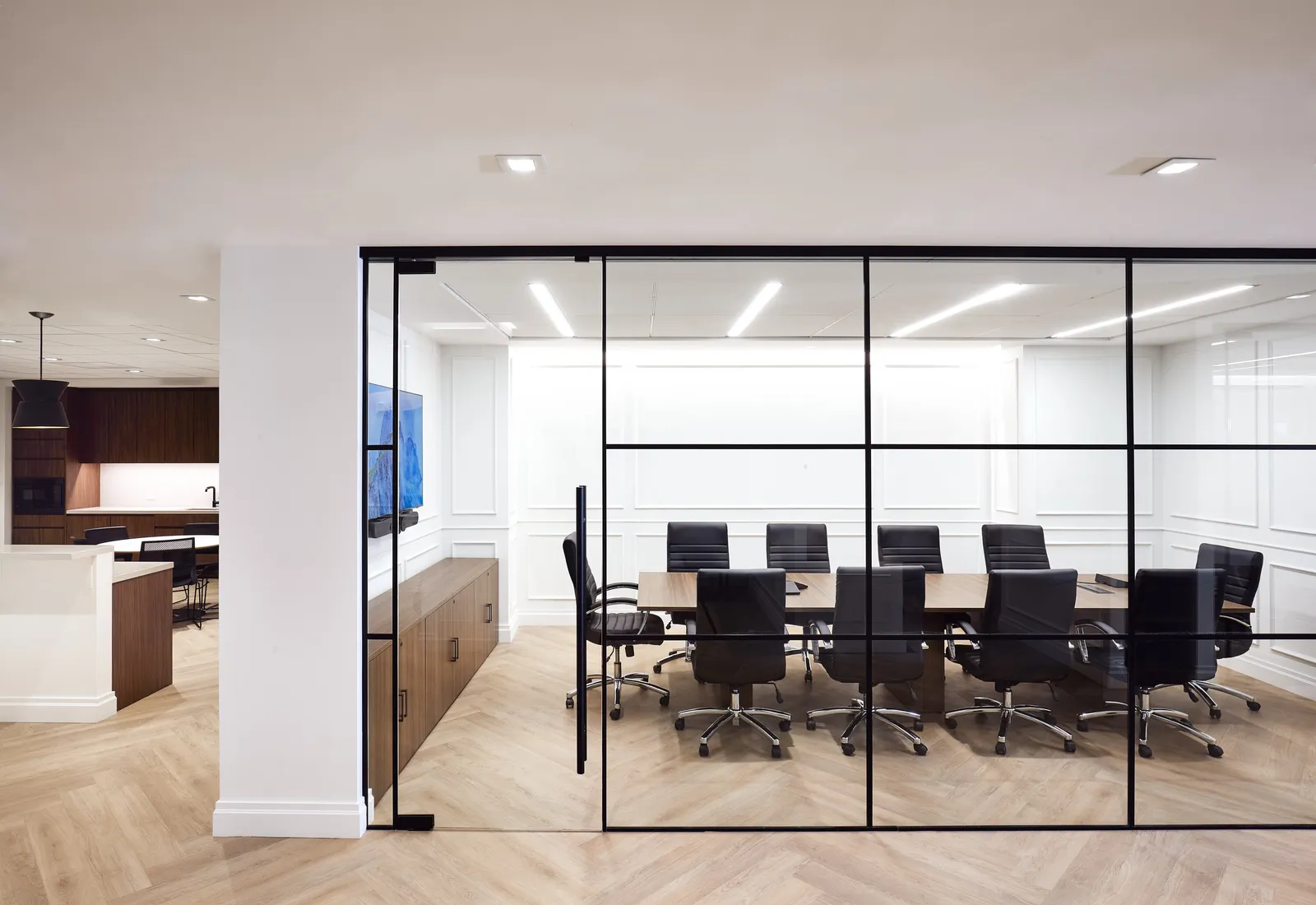 conference room behind a black-framed glass wall, long wood table and black chairs, kitchen at left