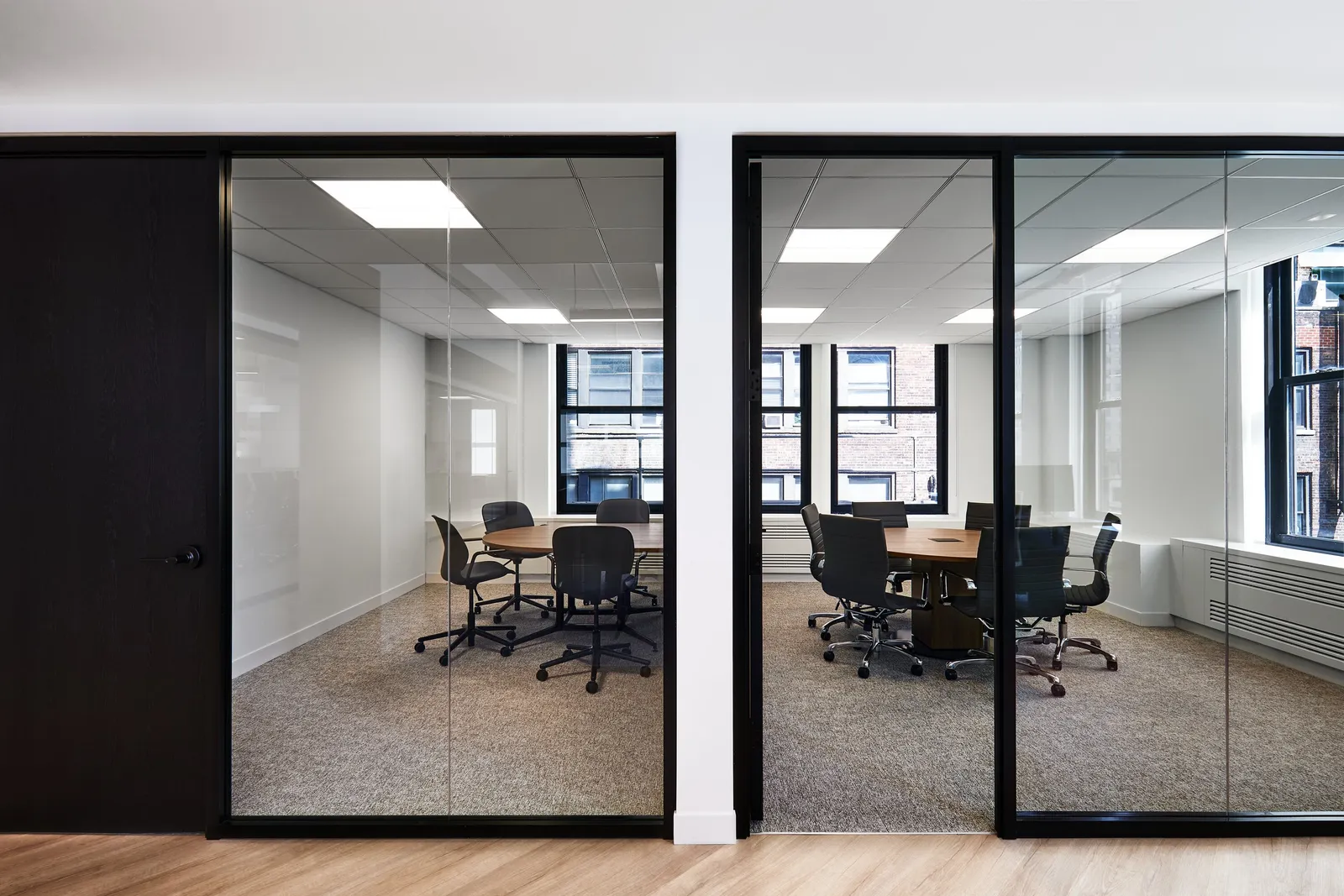 two glass-fronted meeting rooms with black frames, round tables and chairs, wood floor in front