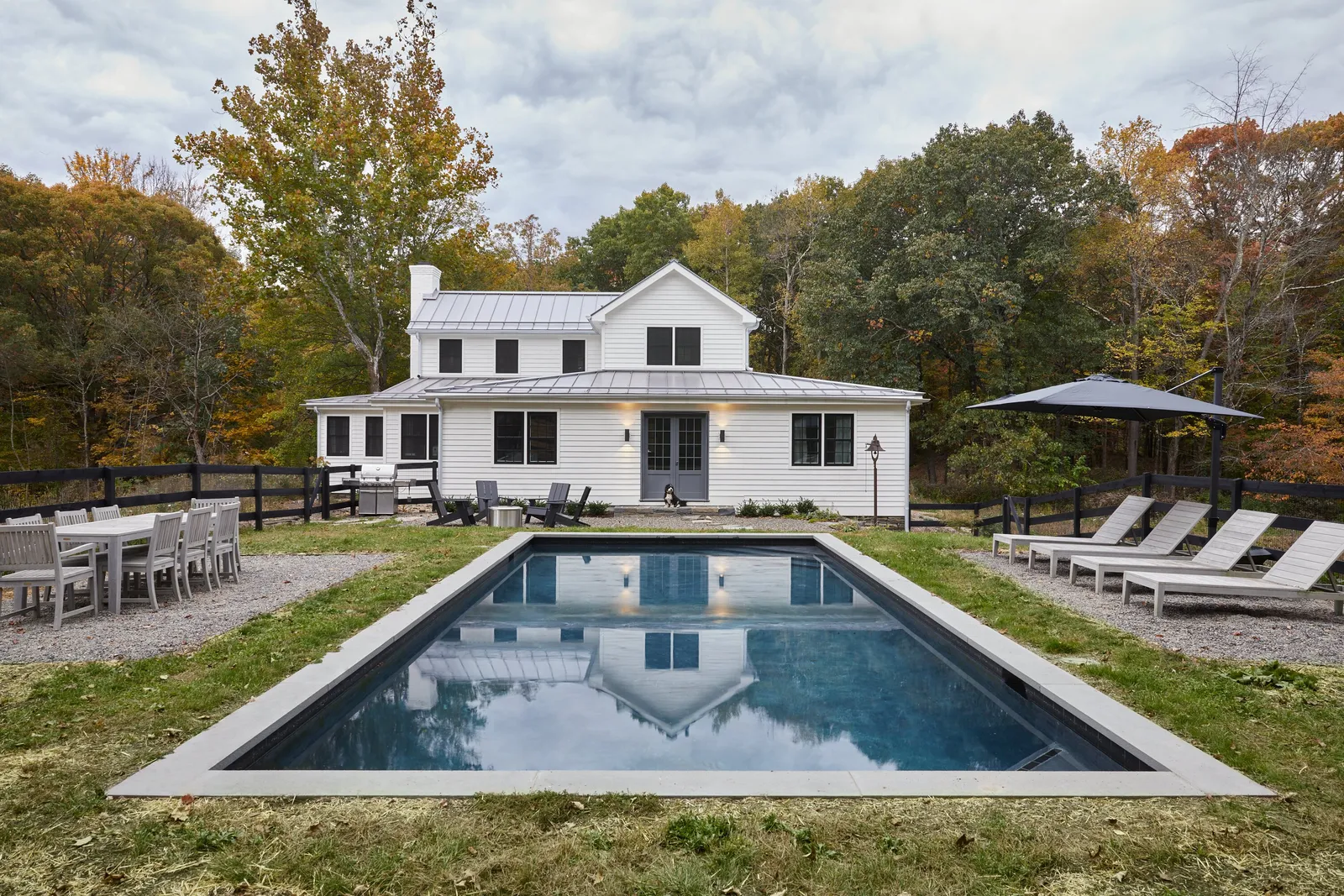 rear of the white farmhouse beyond a rectangular pool, with lounge chairs and a dining table on the lawn