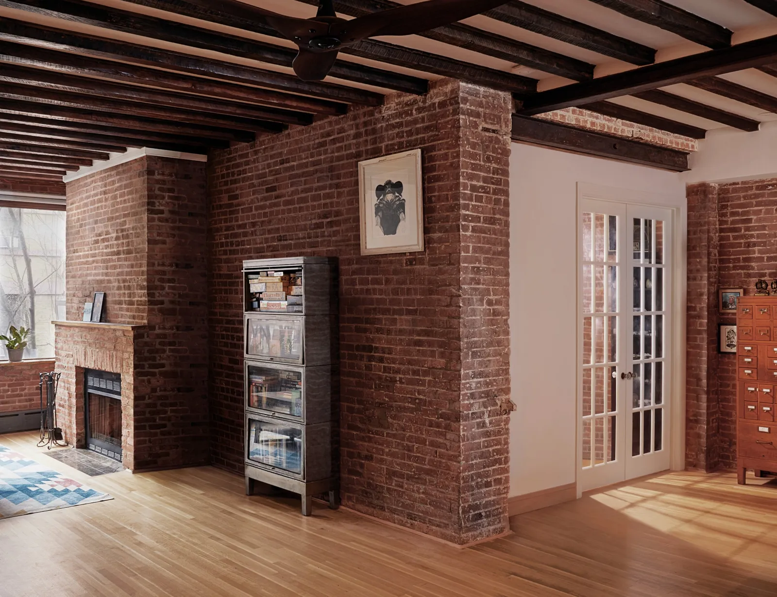 brick-walled room with dark ceiling beams, a fireplace, a glass-front cabinet and french doors on the right