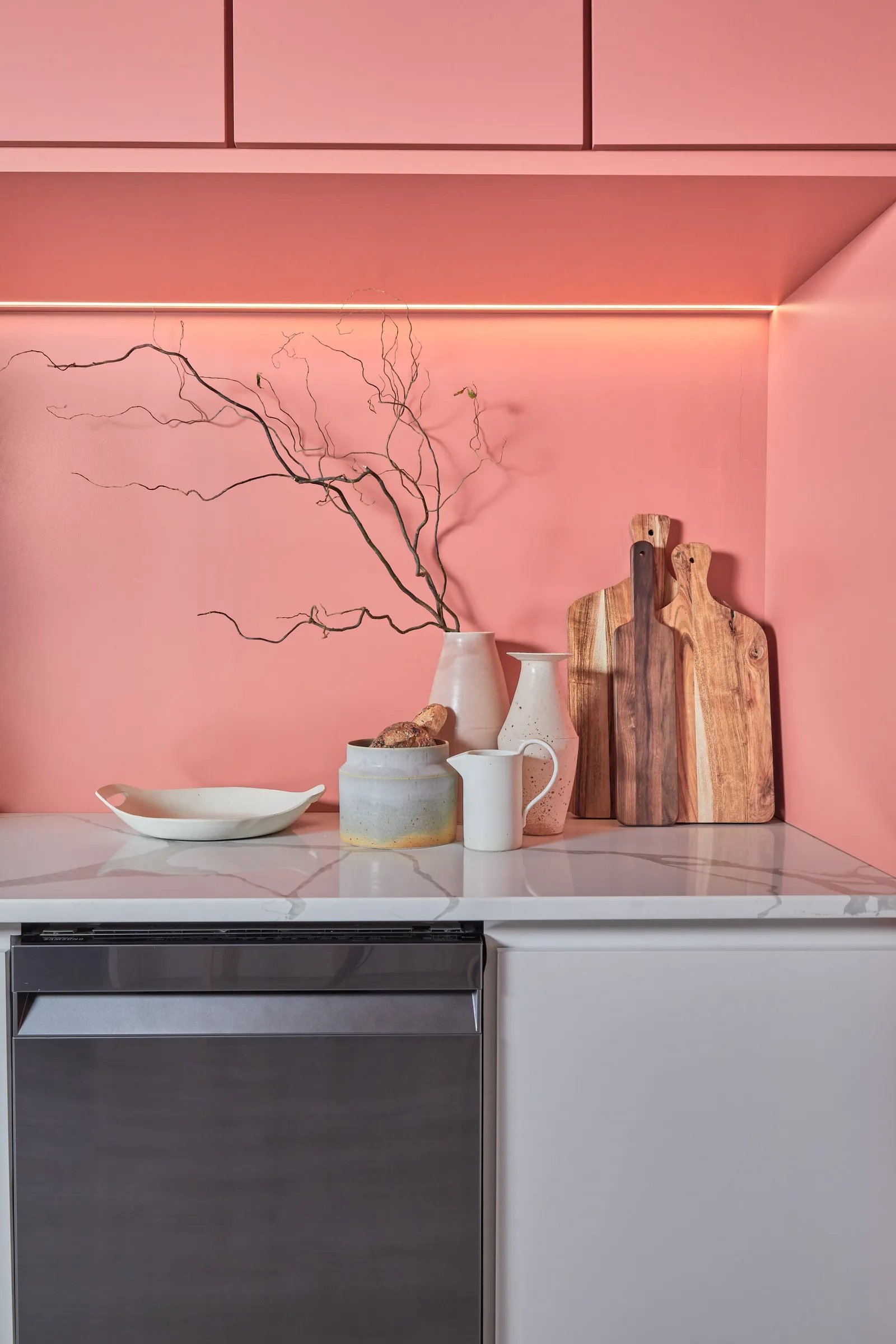 pink backsplash with a branch in a white vase, wooden cutting boards, ceramics and a dishwasher below