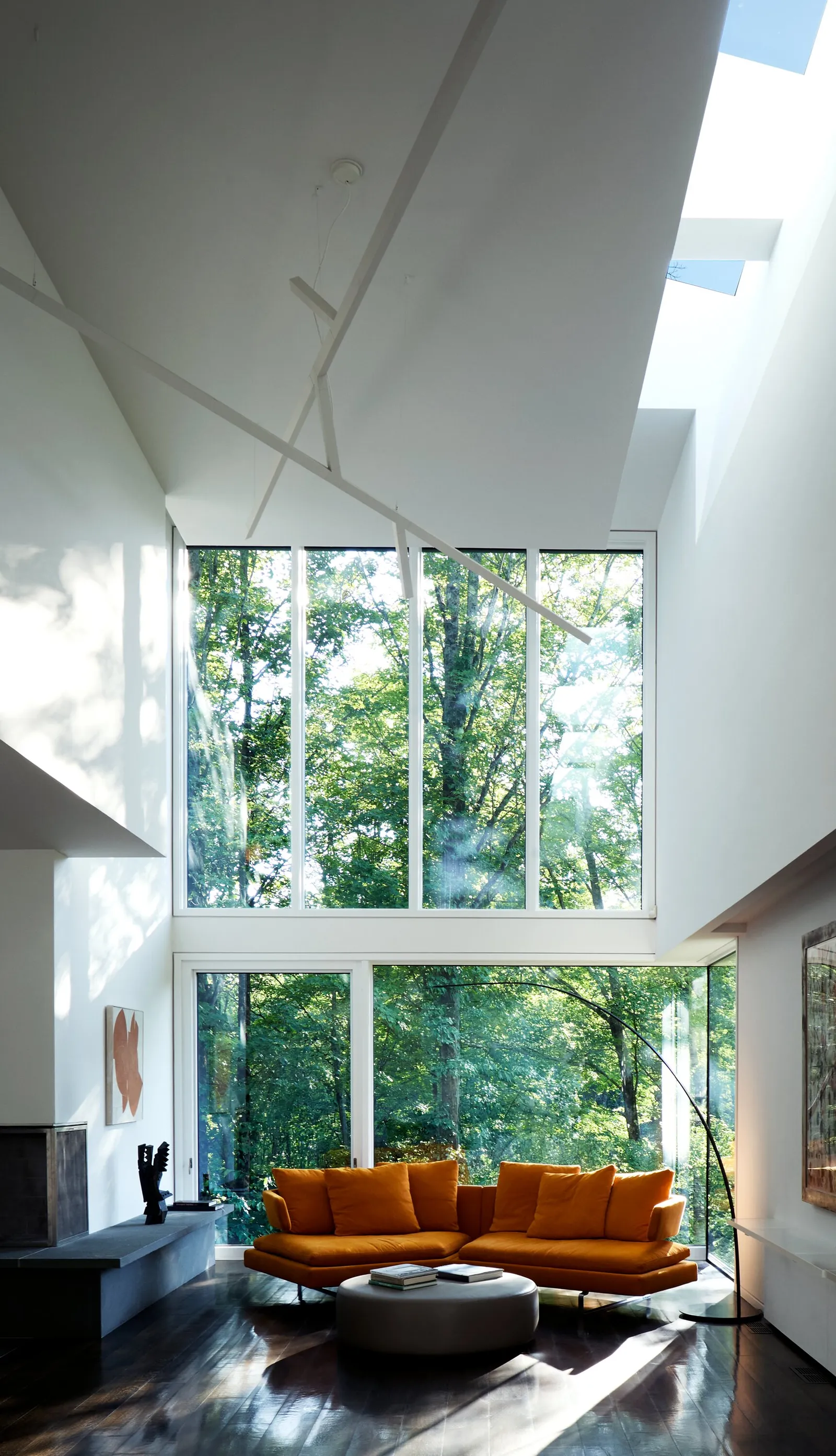 double-height living room with a tall wall of windows to the trees and an orange sofa under diagonal white beams