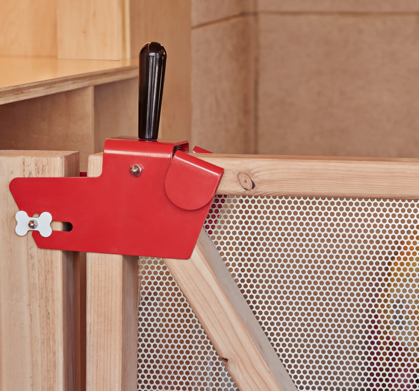 close view of a red metal latch shaped like a dog's head with a bone-shaped pull, on a wood gate with perforated panel
