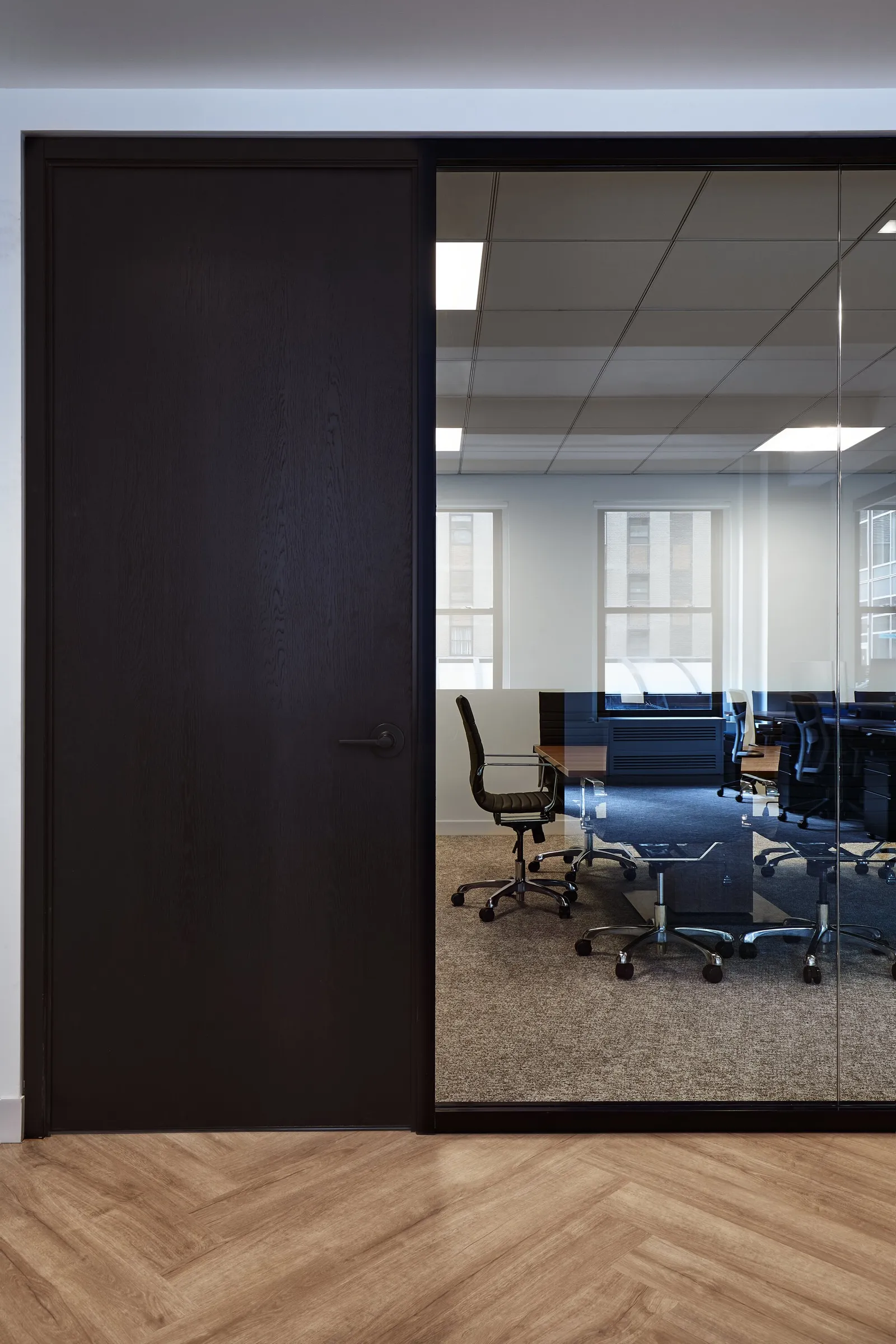 dark wood door beside a glass wall looking into a conference room with navy desks and windows