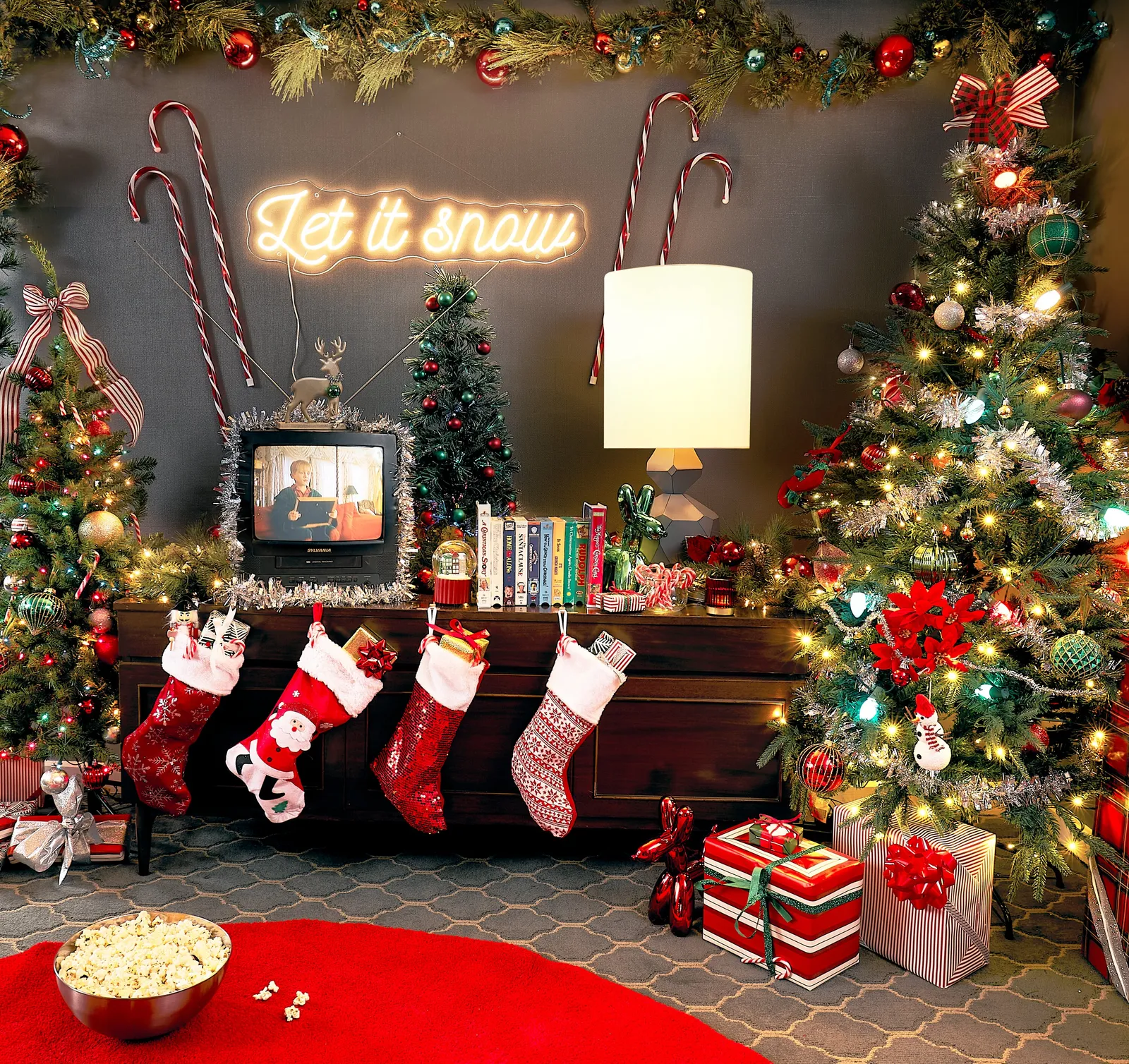 wall of stockings on a dark console under a "let it snow" neon sign, a tinsel-framed tv, and a bowl of popcorn on the red rug