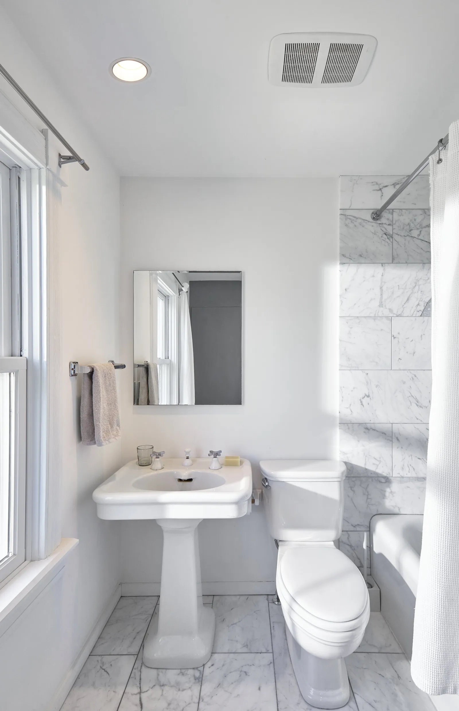 small white bathroom with a pedestal sink, toilet, marble-tiled tub wall and a mirror