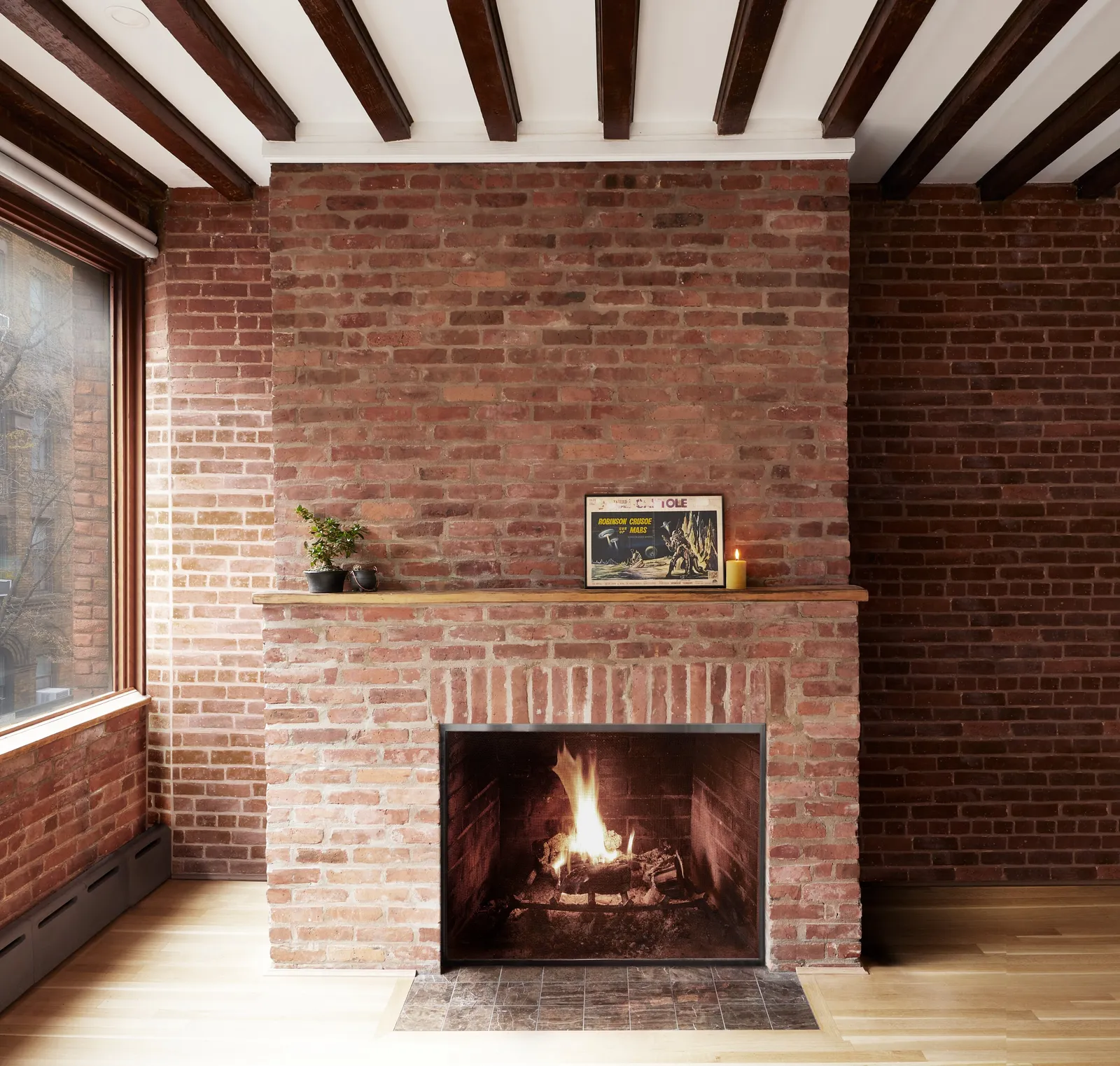brick fireplace with a lit fire, wood mantel, framed poster and a candle, under white ceiling with dark beams