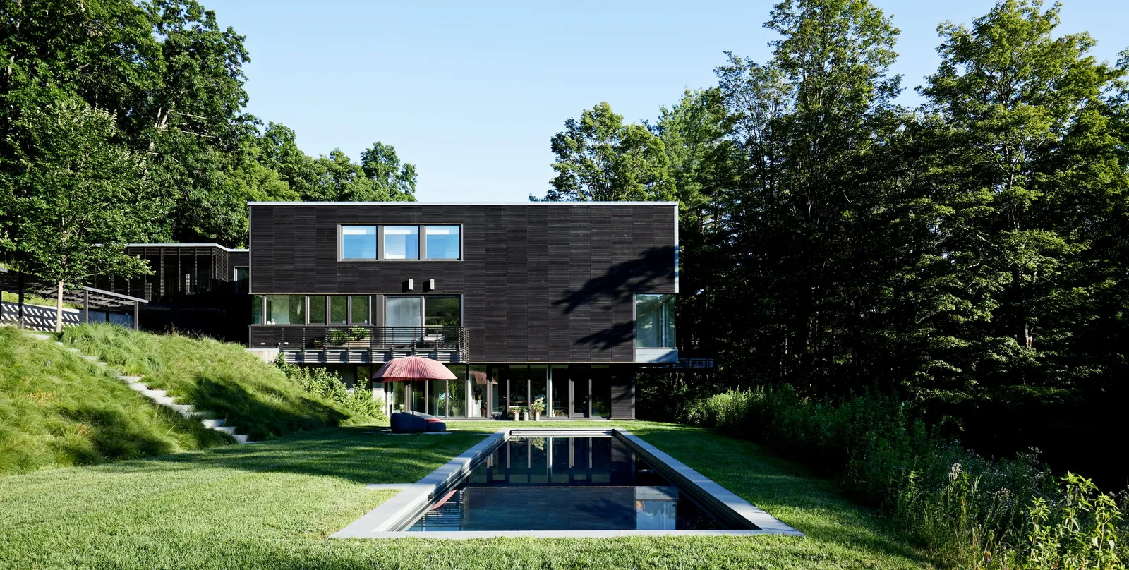 dark wood-clad modern house across a lawn with a long reflecting pool, steps and a hillside to the left