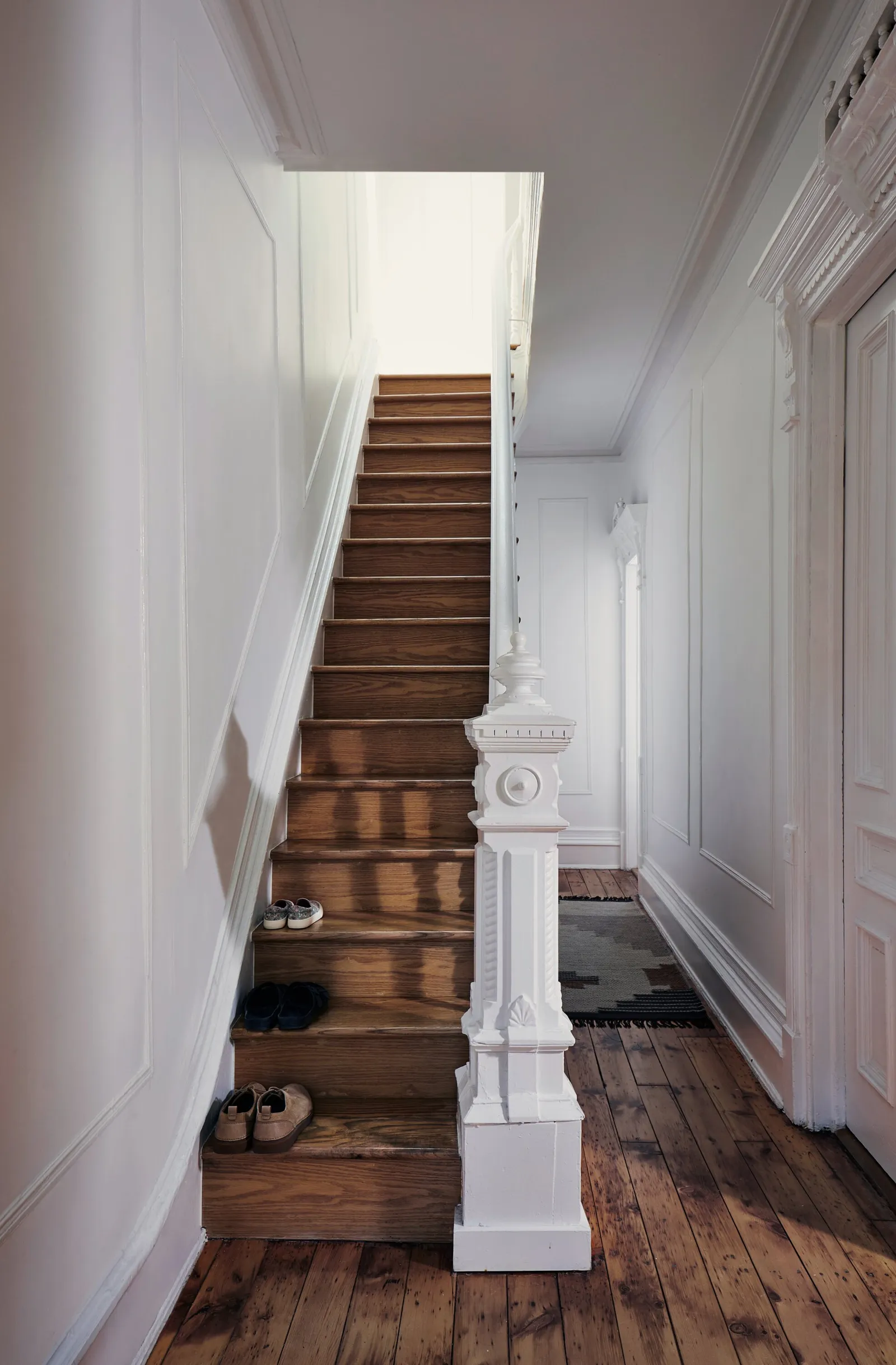 narrow wooden staircase with a white carved newel post, shoes on the steps and worn plank floors