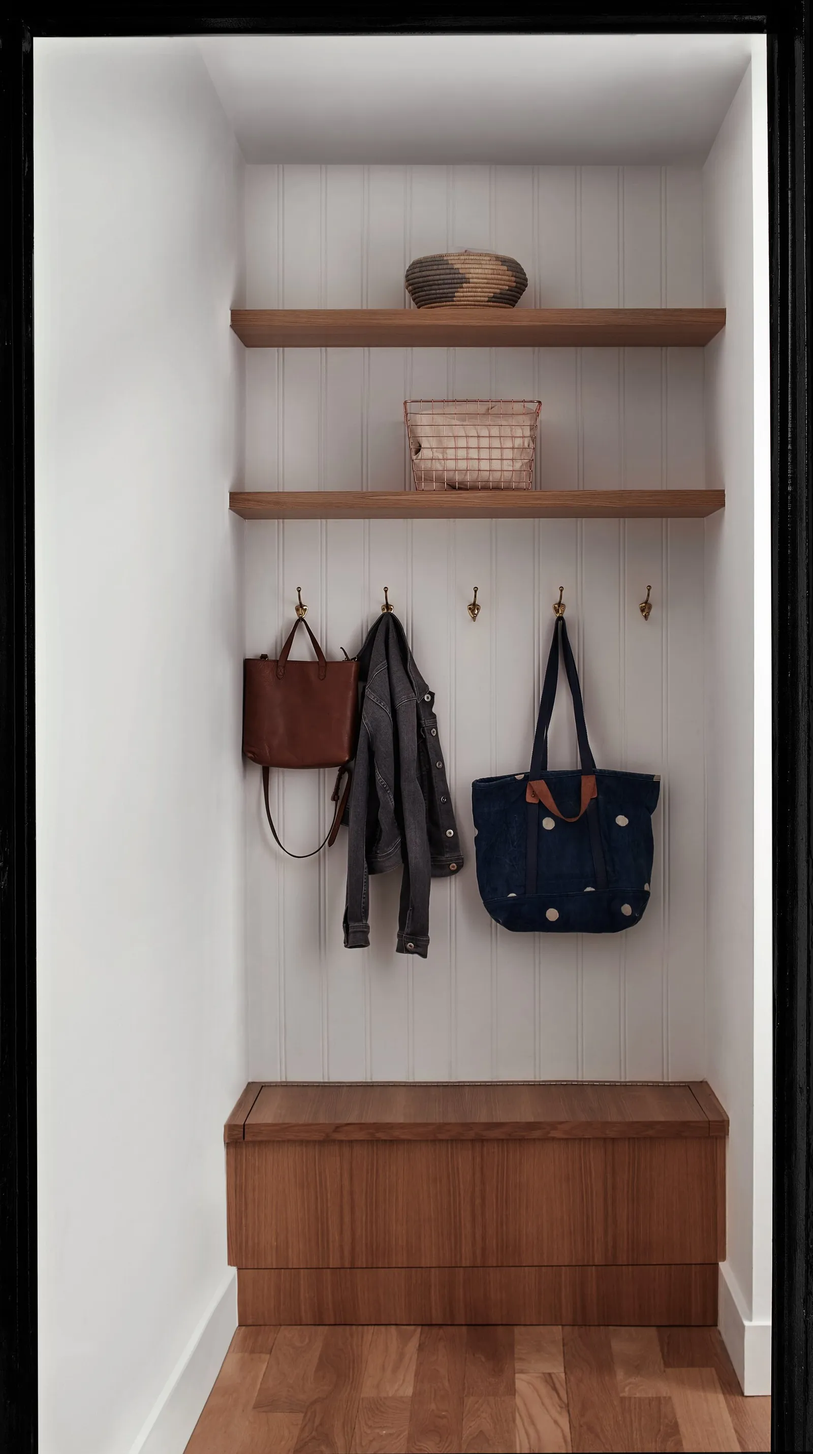 narrow entry nook with wooden shelves, brass hooks, a bench and a bag and jacket hanging on the wall