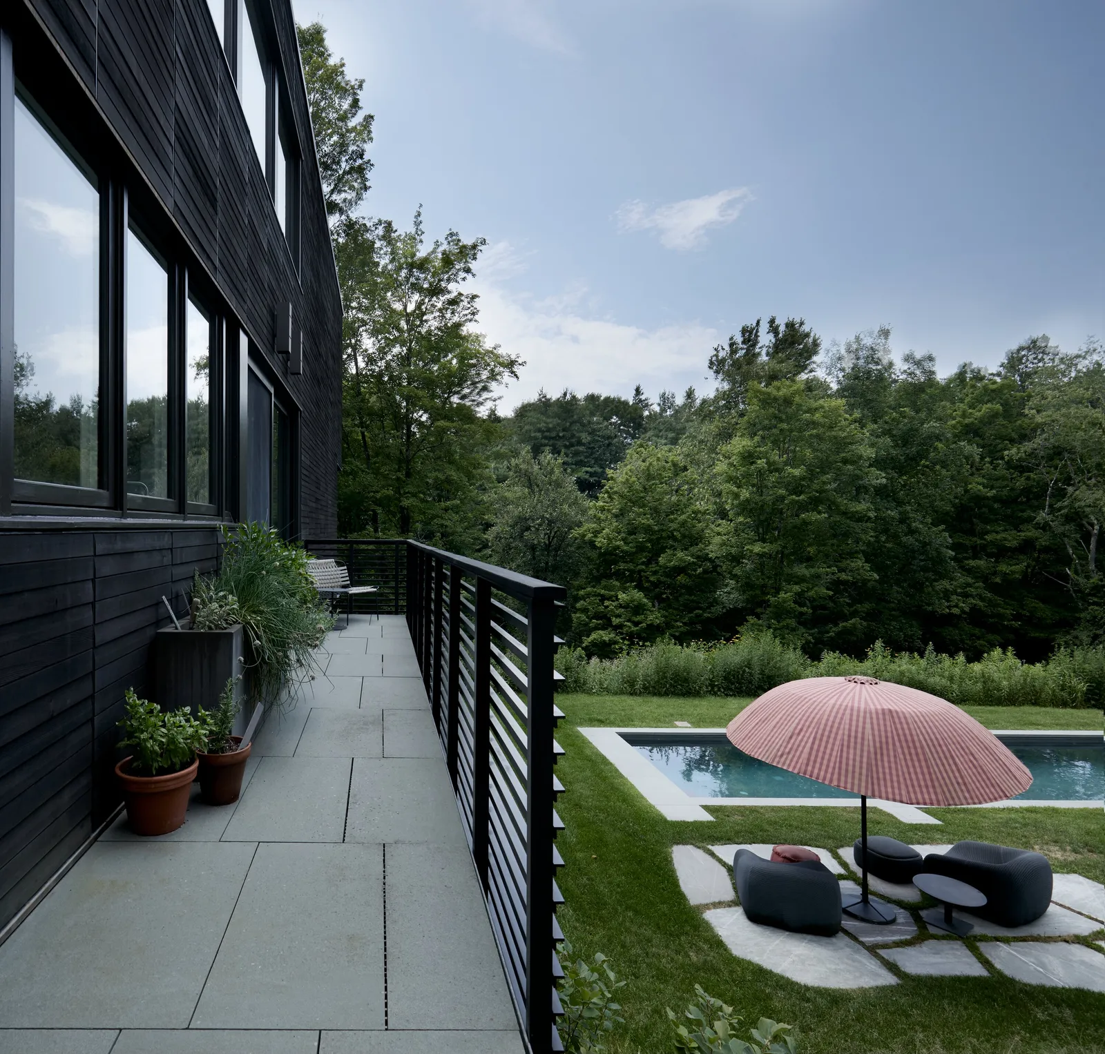 stone terrace with a black slatted railing beside the dark house, looking over a pool and umbrella to woods