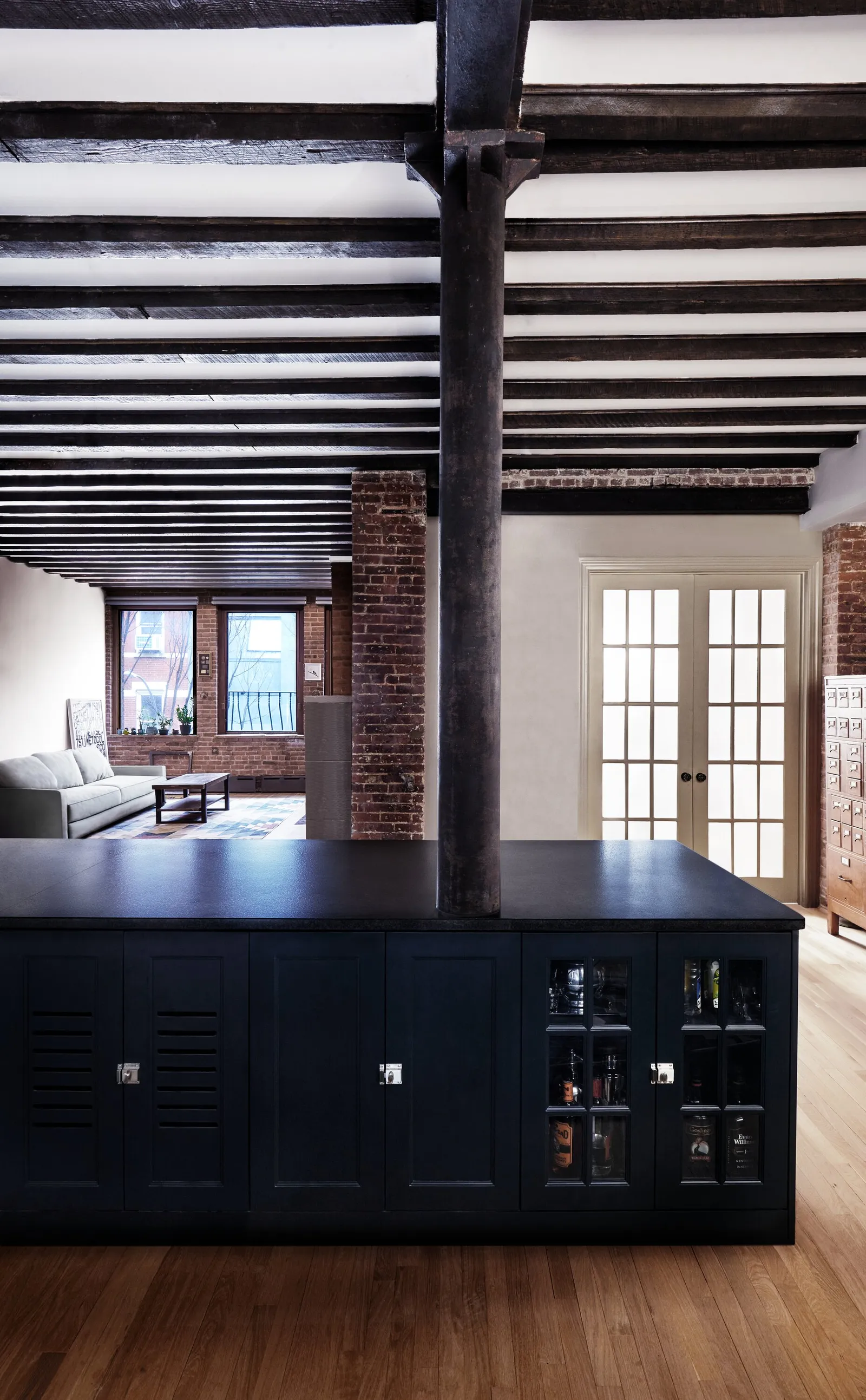 long loft view past a dark cabinet island and a black iron column to a sofa and brick wall under dark beams