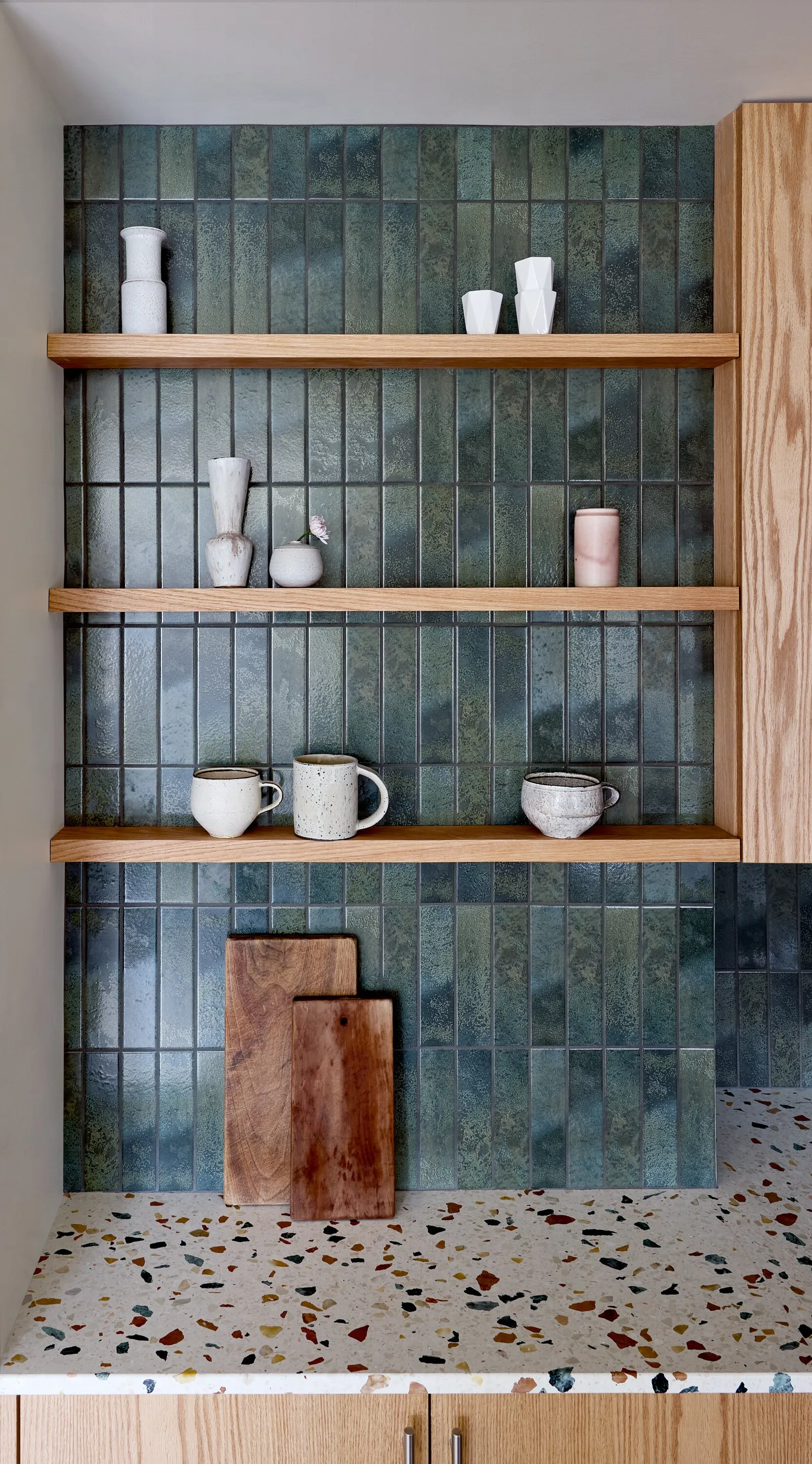 open shelves on blue-green tile with mugs, vases and cutting boards above a terrazzo counter