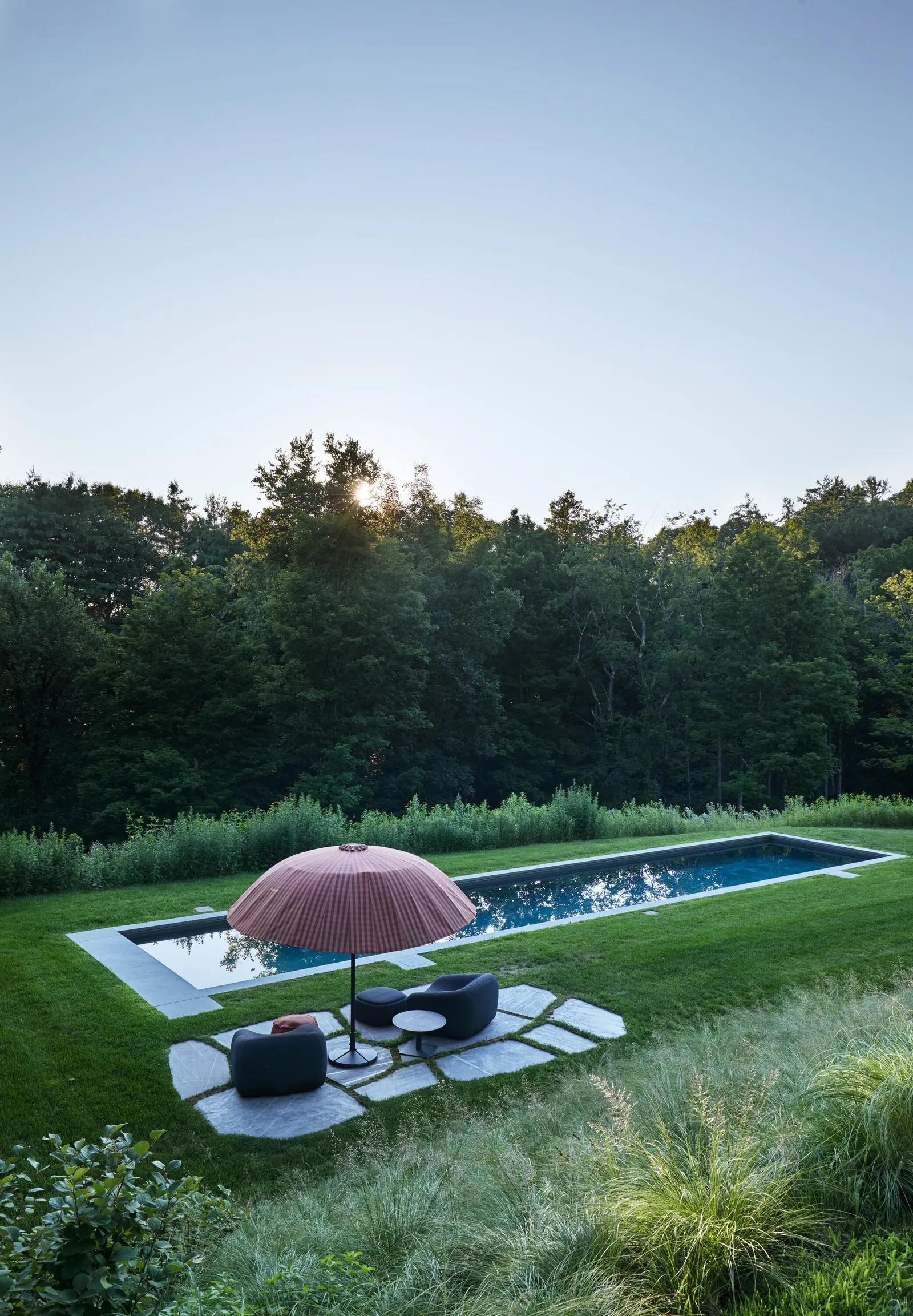 high view of a long narrow pool and pink umbrella with lounge chairs on stone pads, backed by dense forest