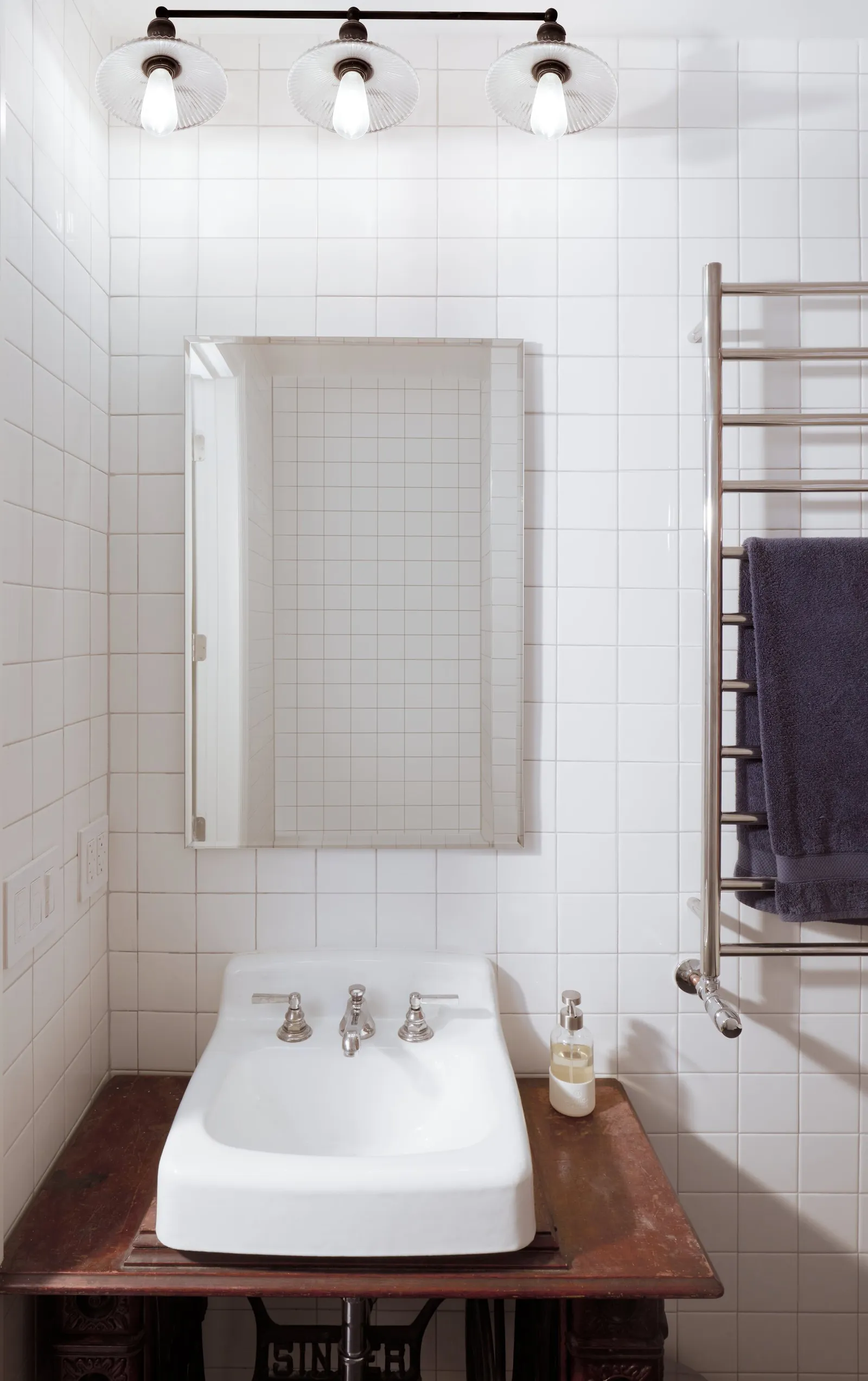 small bathroom with white square tile, a wall-mounted white sink on a wooden table base and a towel warmer