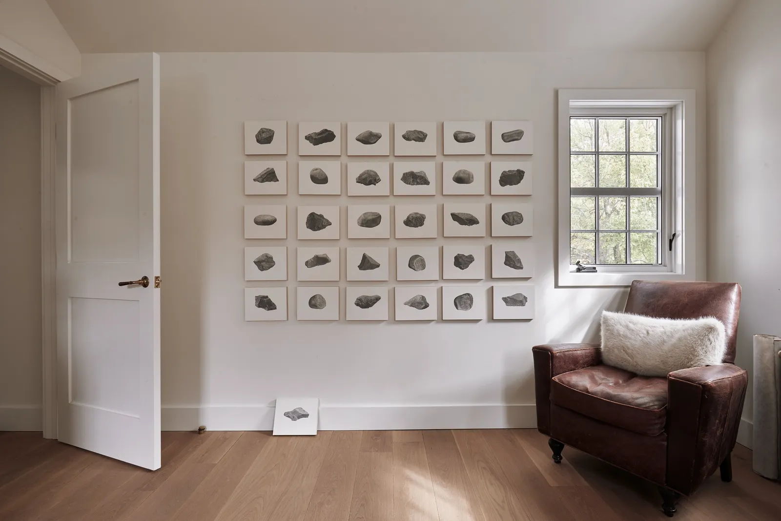 wall of small framed stone drawings beside a window and a leather armchair, with a white paneled door at left