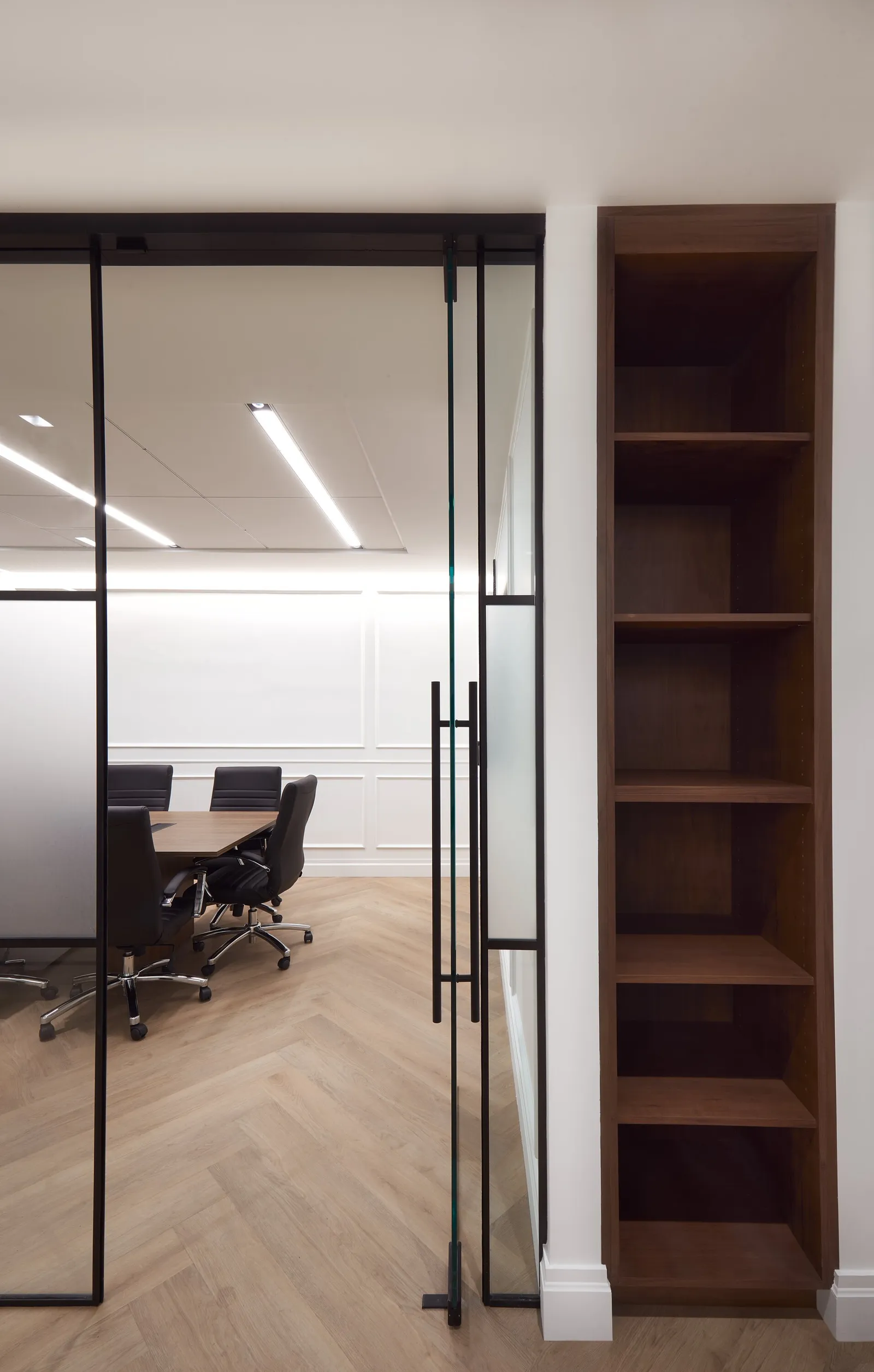 glass door into a conference room beside a tall walnut open bookshelf