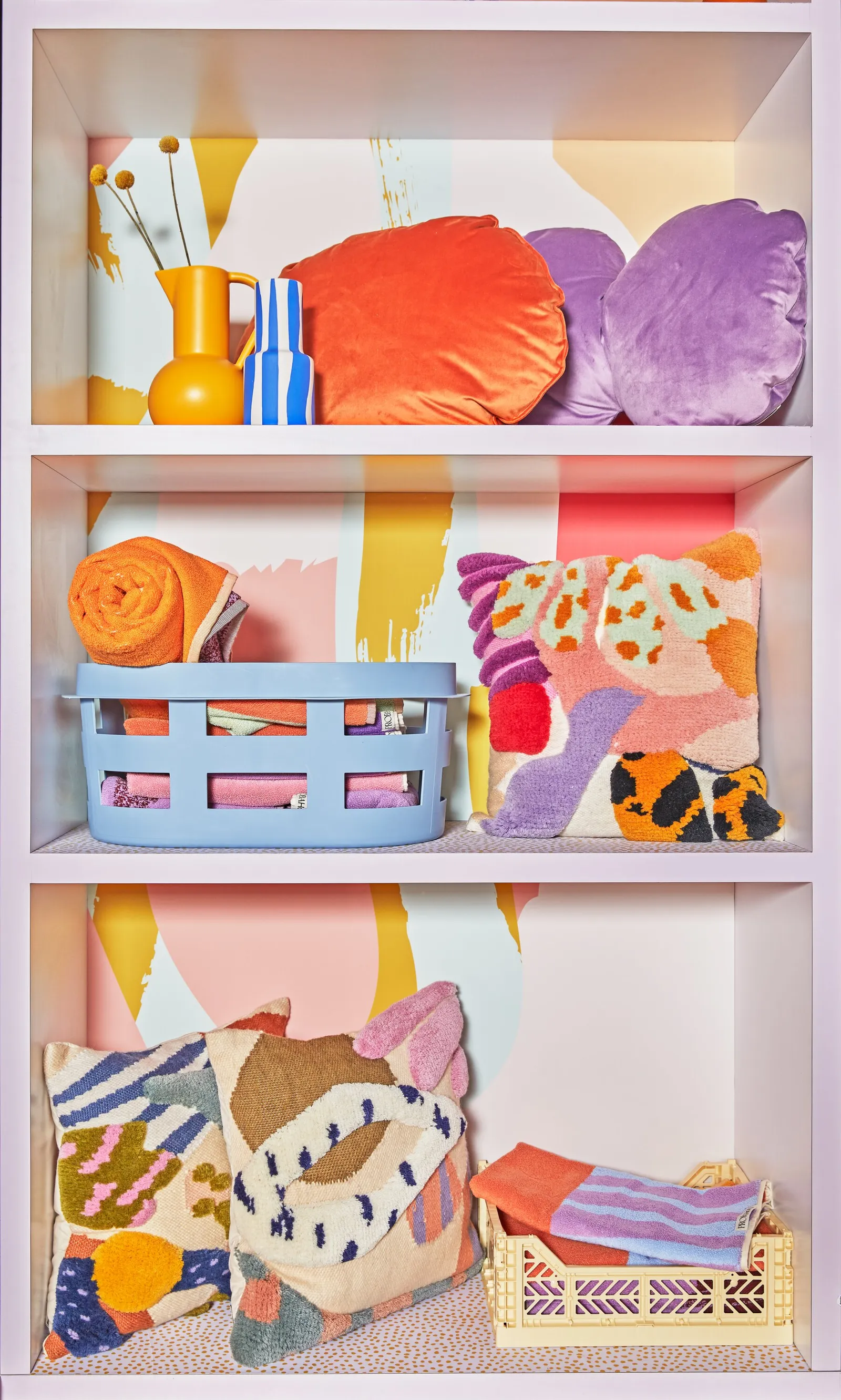 shelves holding orange and purple velvet cushions, yellow vase, blue laundry basket and folded towels