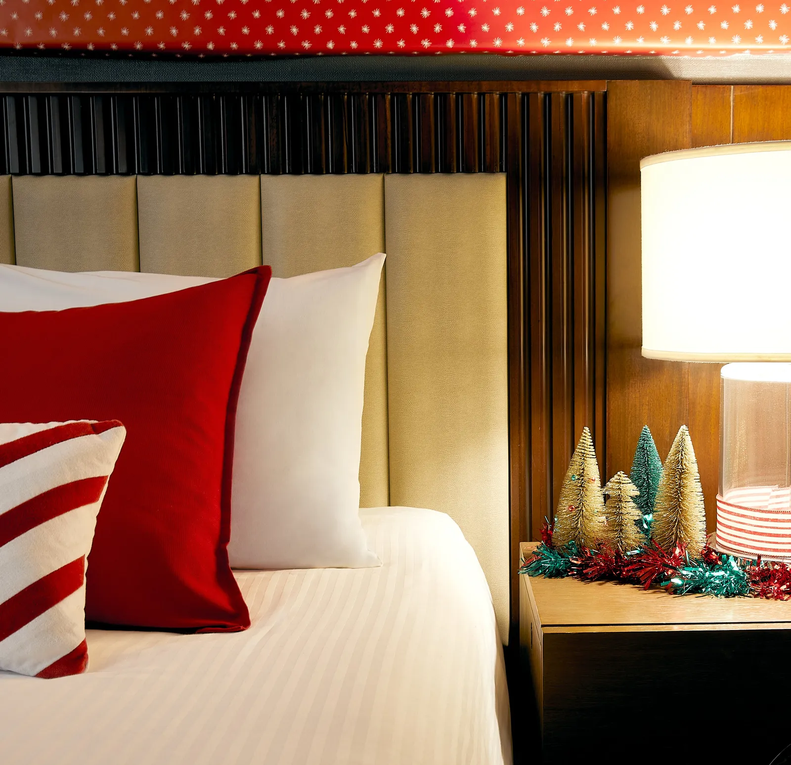 close view of a bed's tan paneled headboard, red and striped pillows and a nightstand with small gold bottle-brush trees