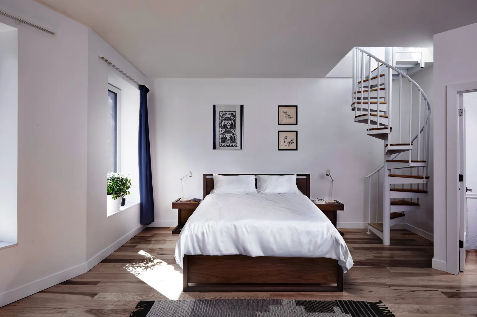 bedroom with a white-covered wooden bed, a white spiral staircase in the corner and a navy curtain at the window