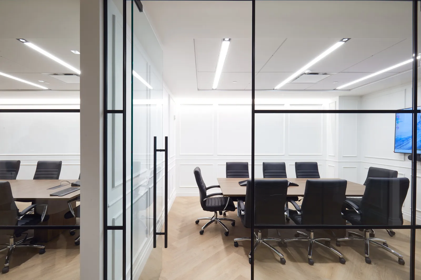 conference room through black-framed glass panels, black chairs around table, wall screen at right