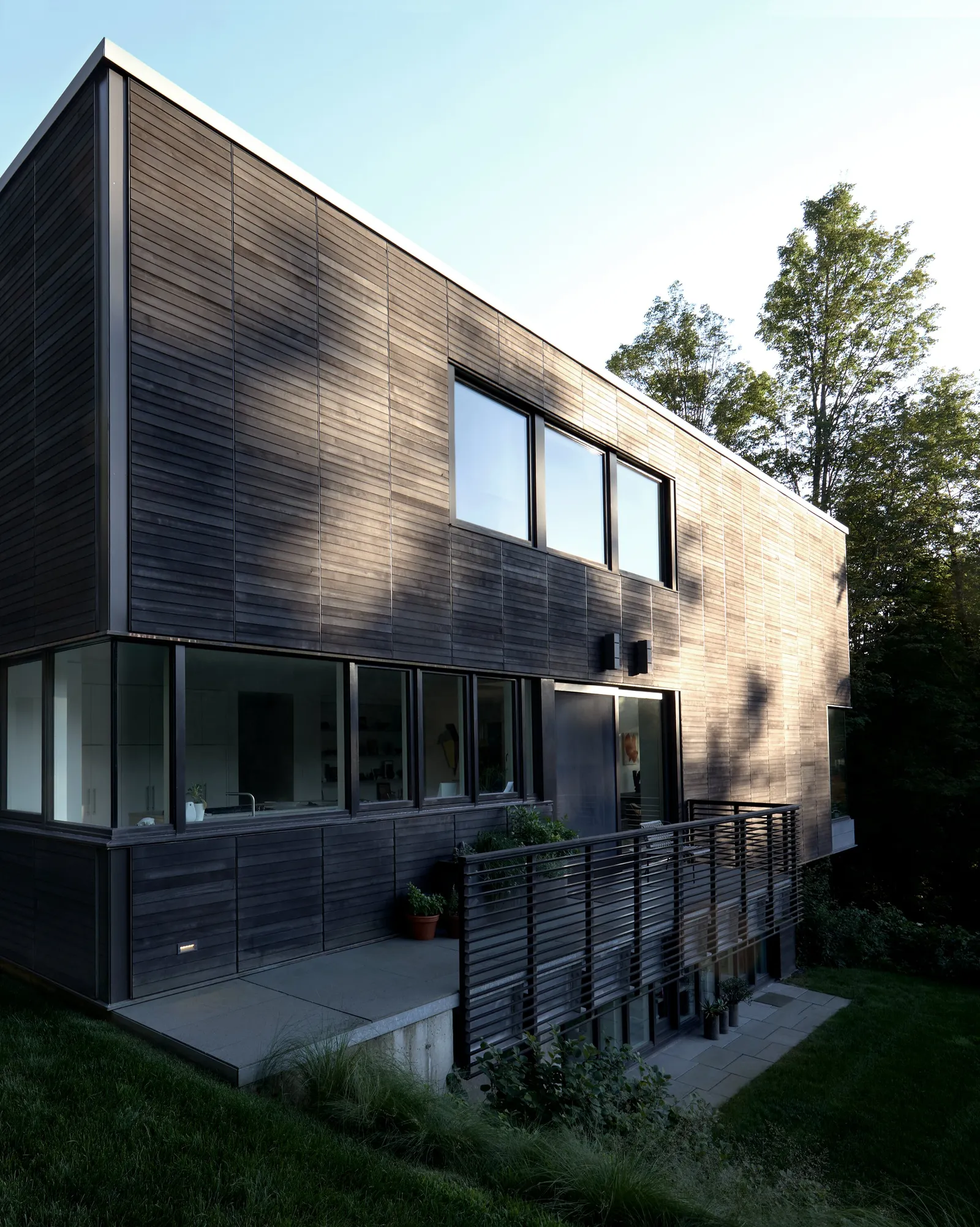 corner of the dark horizontal-board house with large windows, a deck rail and grass in the foreground