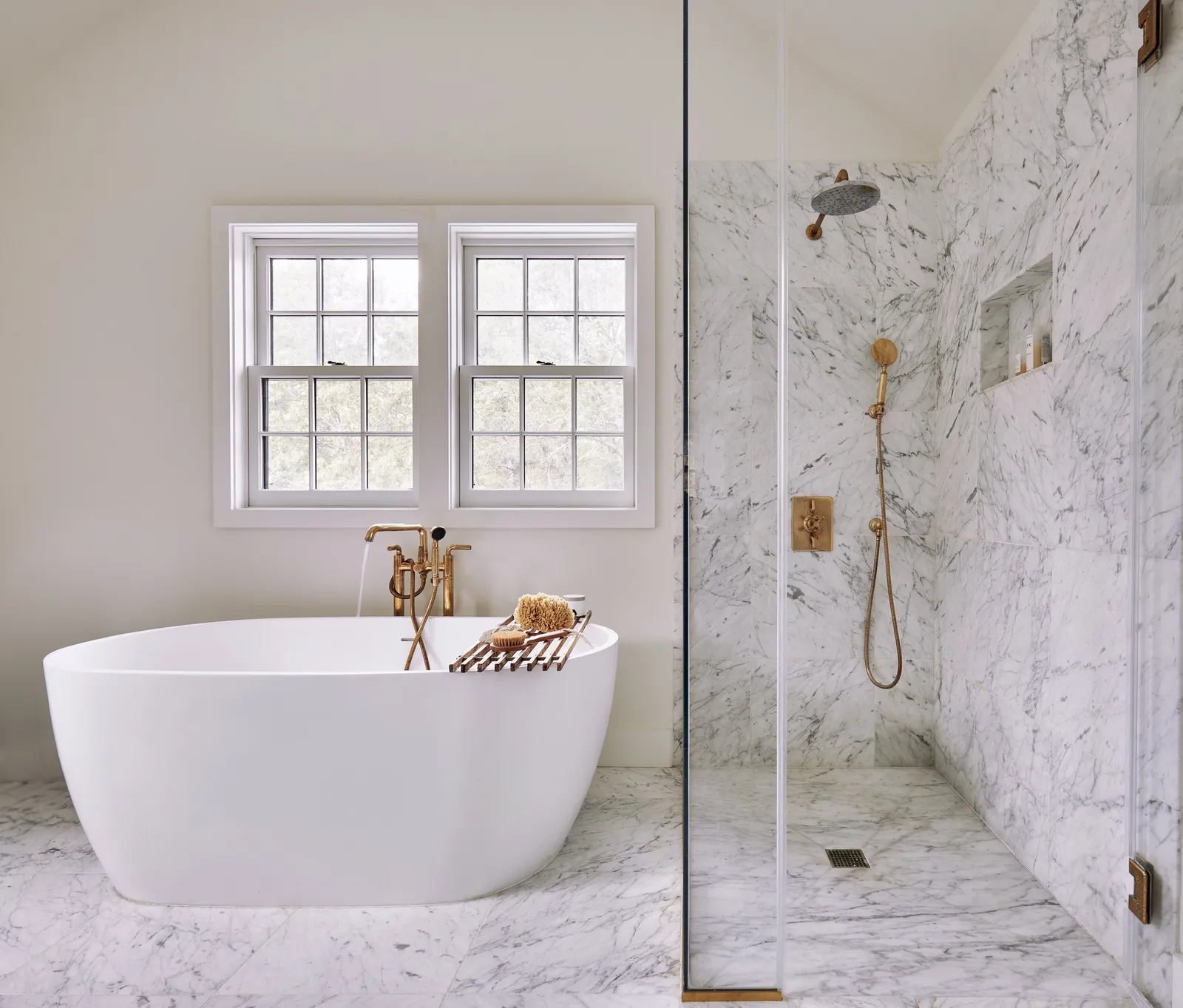 freestanding white tub with brass fittings beside a glass marble shower under a pair of windows