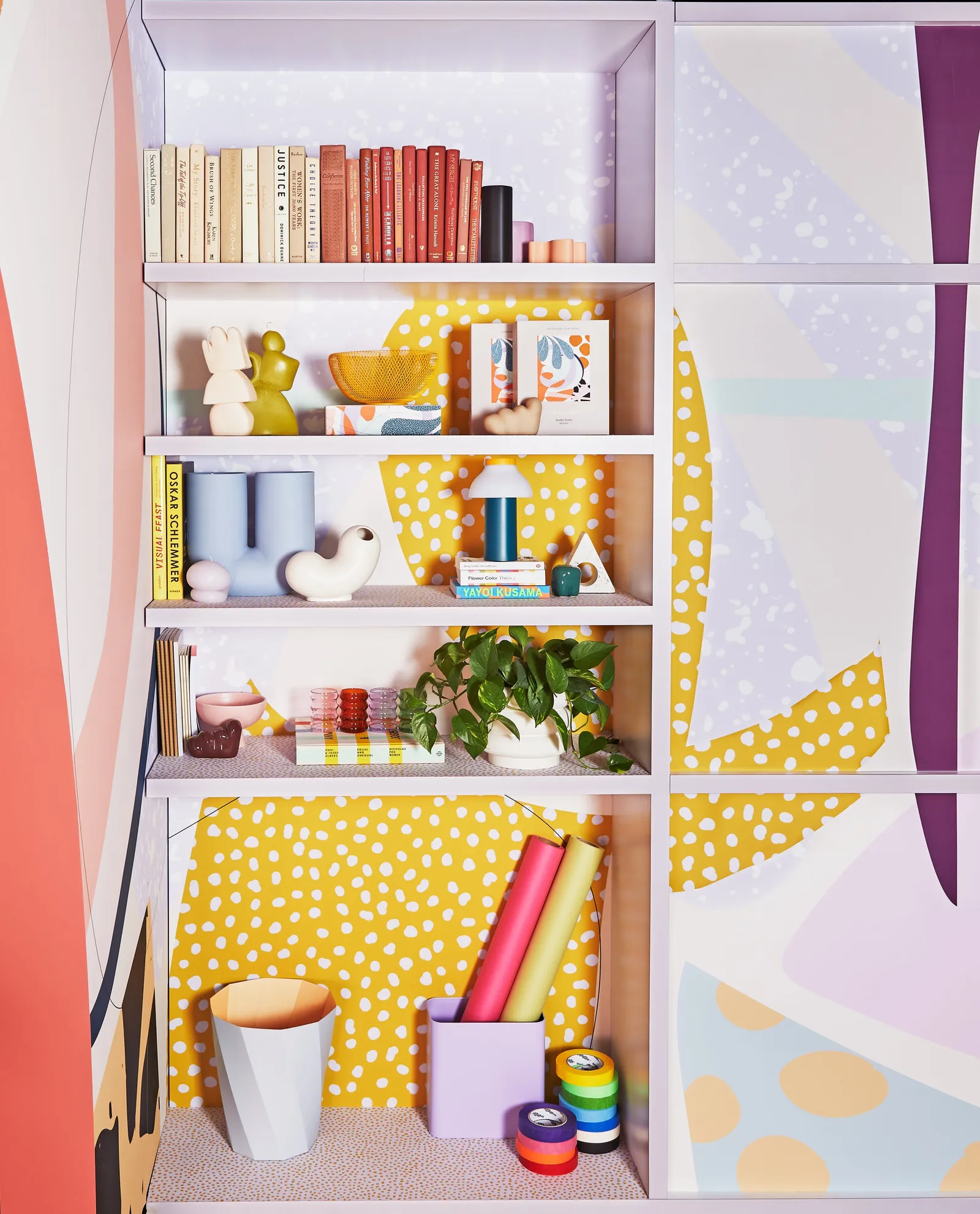 bookshelf with colorful books, small sculptures, potted plant, wrapping paper rolls and tape on dotted yellow wall
