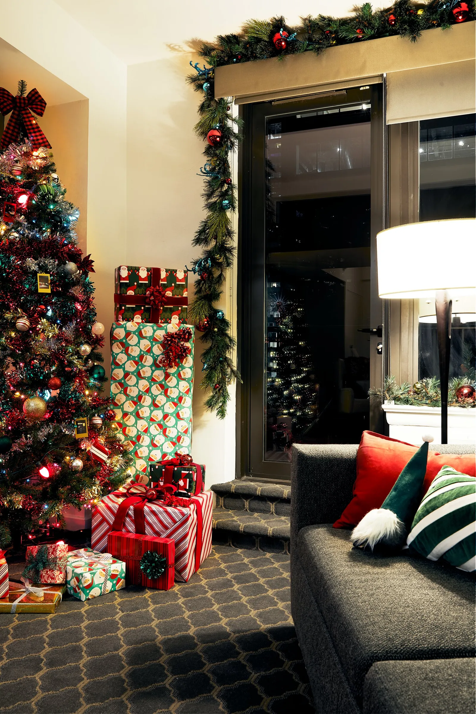 vertical view of a tree and stacked wrapped gifts beside a balcony door, with a grey sofa and striped pillows