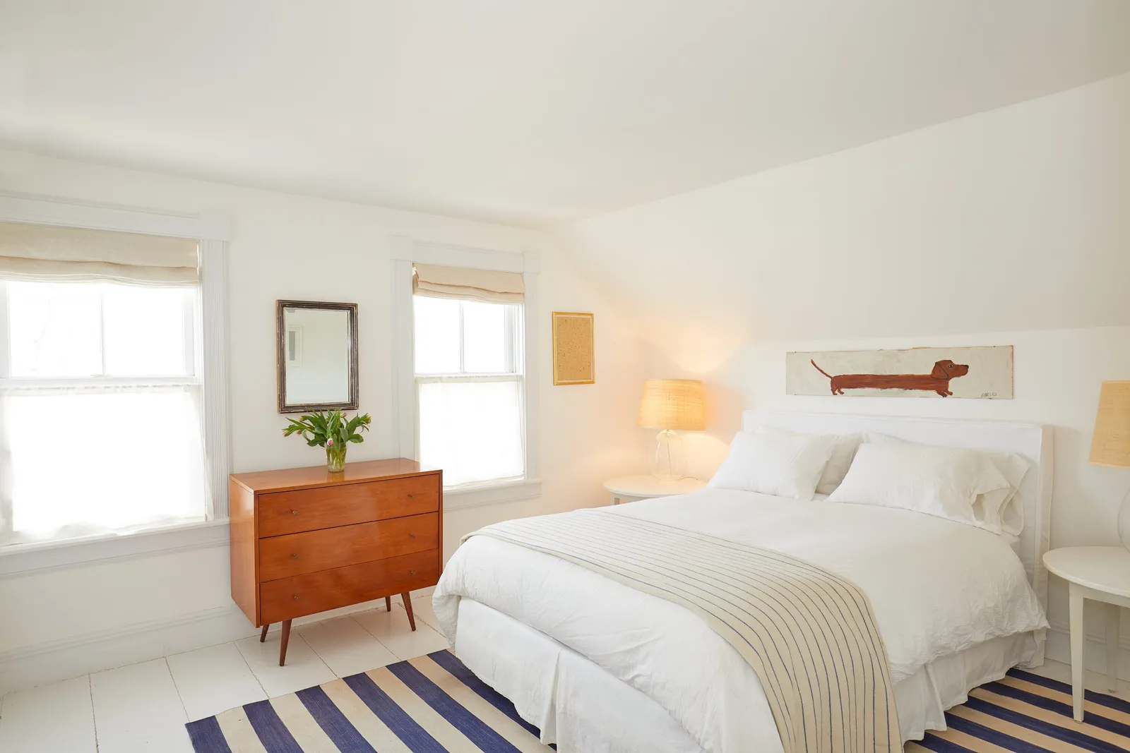 bedroom with a striped blue-and-cream rug, a wooden dresser, a white bed and a dachshund artwork above the headboard