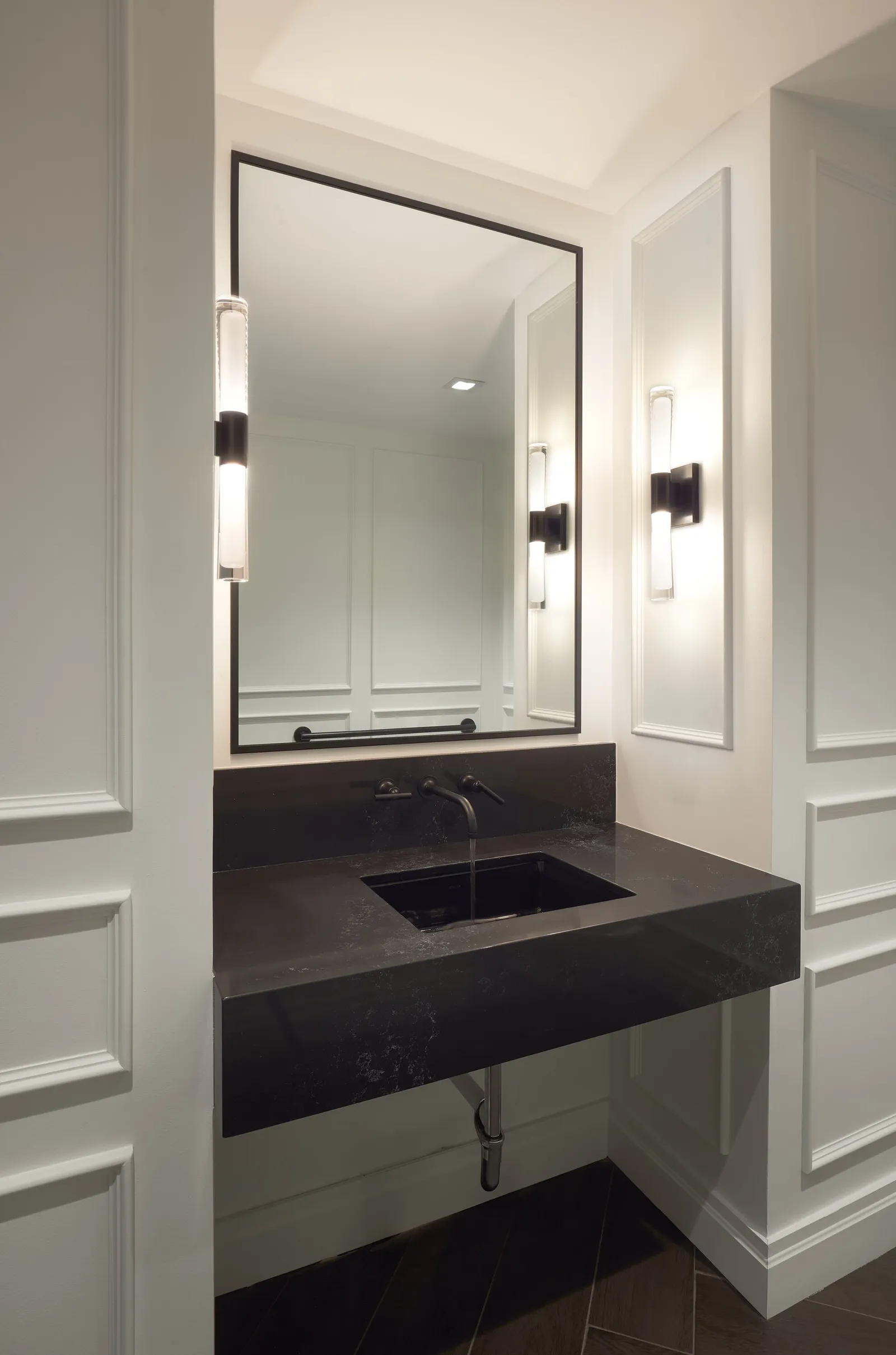 powder room with black stone floating sink, framed mirror and two lit wall sconces on paneled walls