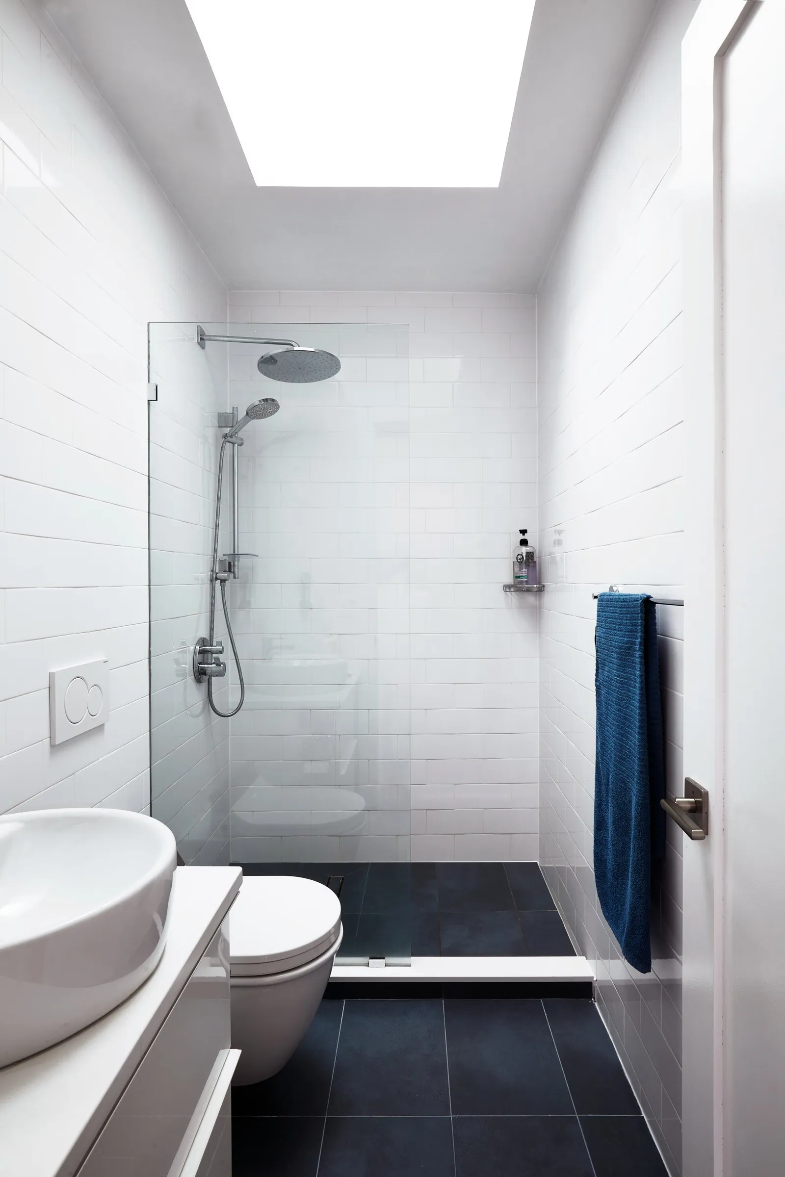 narrow white tiled bathroom with a skylight, glass-walled shower, wall-hung toilet and a blue towel