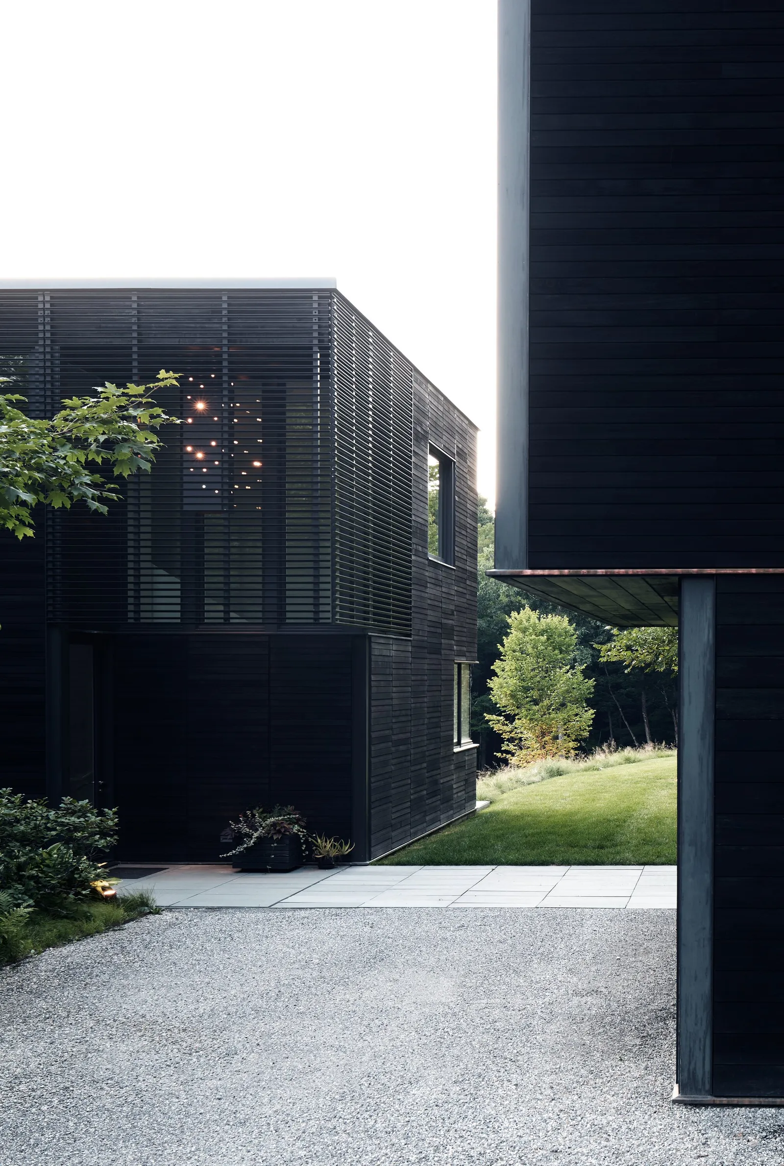 louvered dark wood volume beside a gravel drive, with a light fixture glowing inside and a lawn beyond