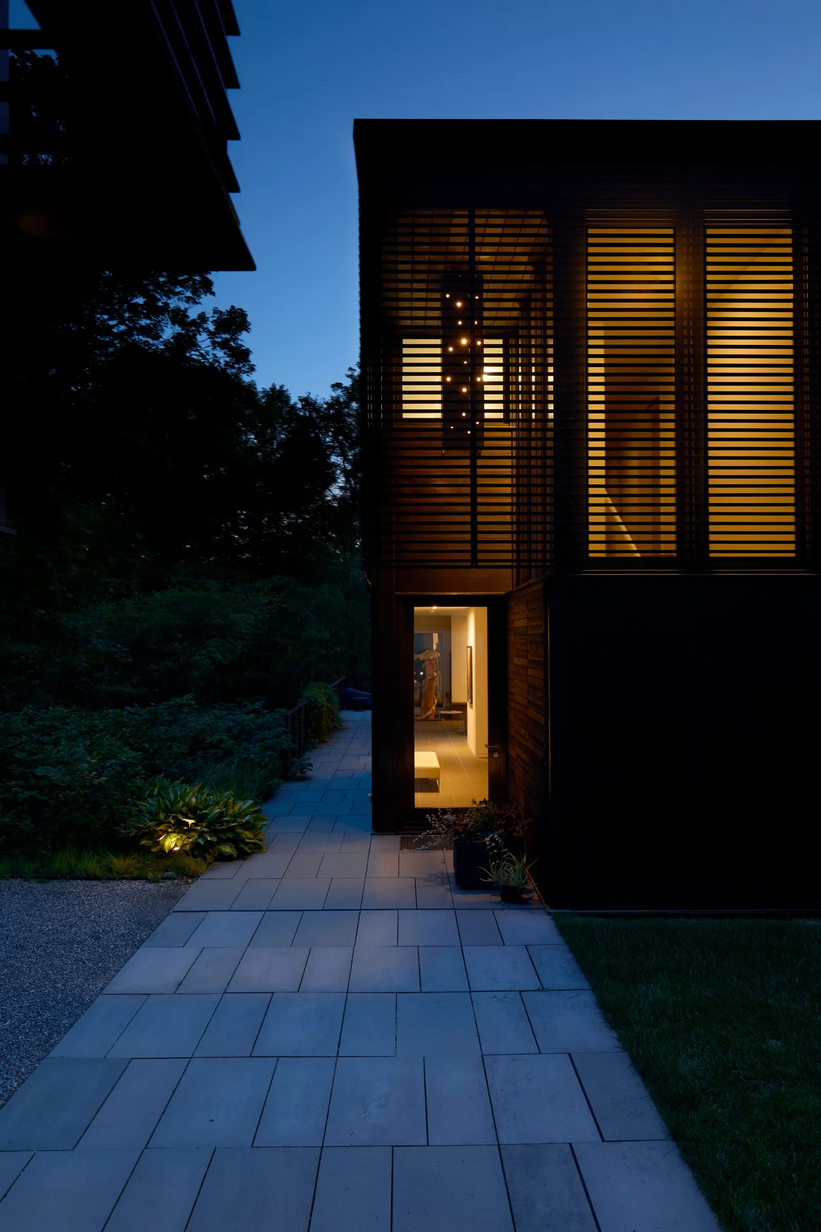 stone path at dusk leading to a glowing entry in a louvered dark wood volume, lit plants along the way