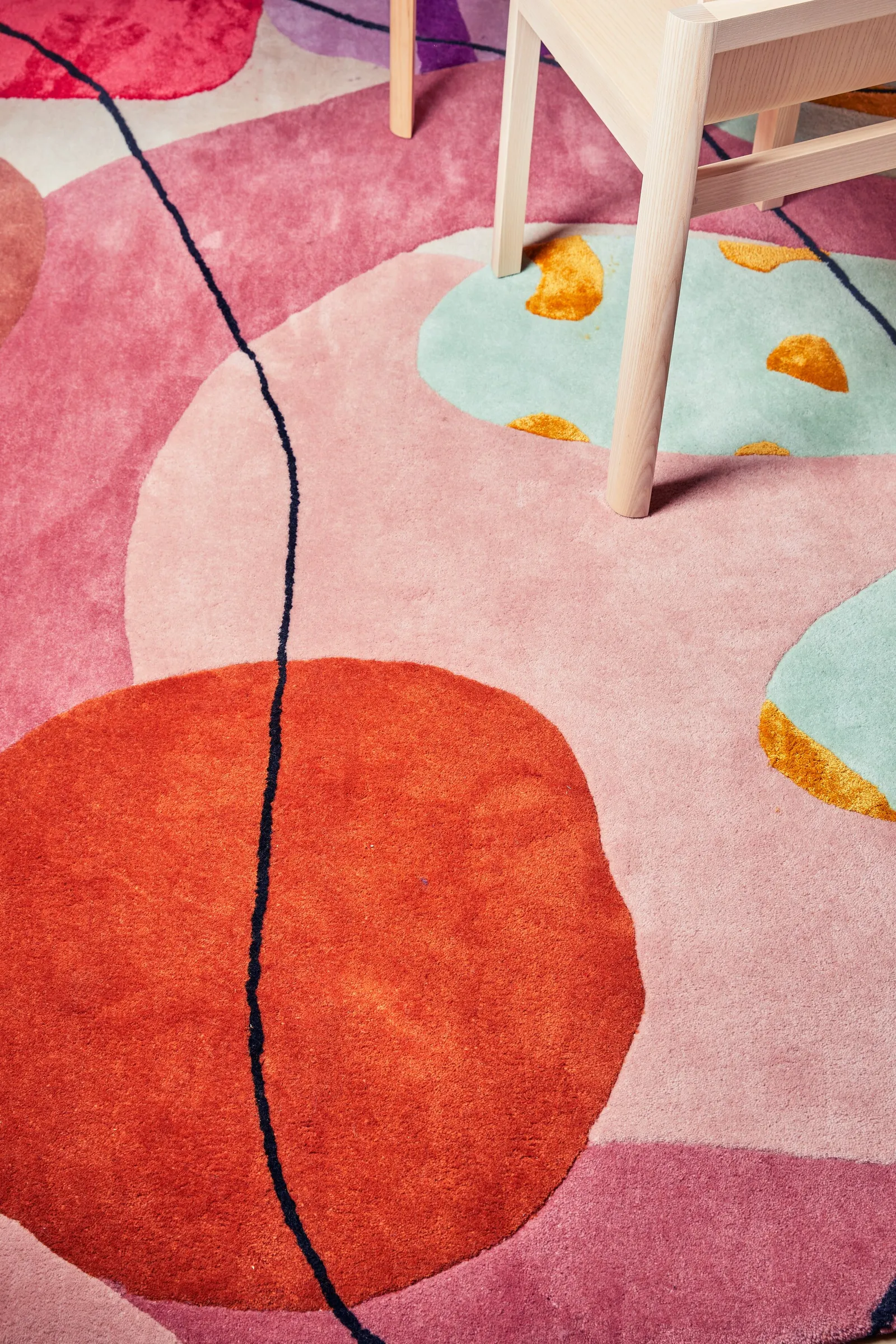 top-down detail of a round rug with red-orange, pink and mint shapes and a dark line, wood chair leg
