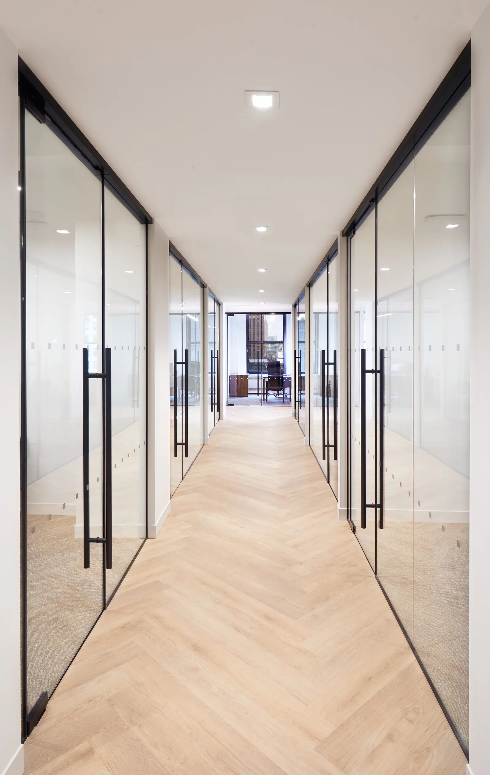 narrow view down a herringbone corridor lined with glass office doors toward a far window