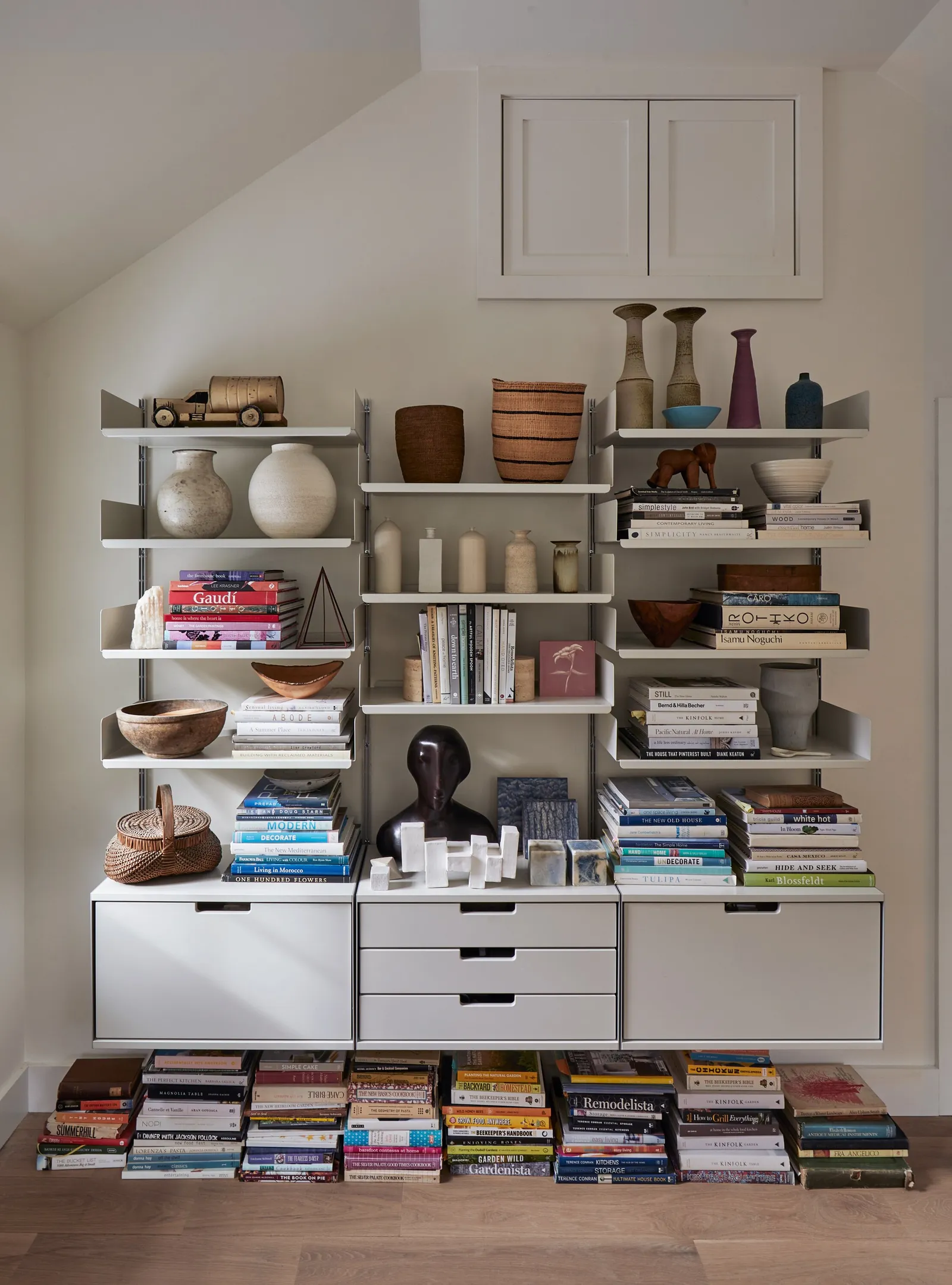 white wall shelving under a sloped ceiling holding ceramics, books and wooden bowls, with more books stacked on the floor
