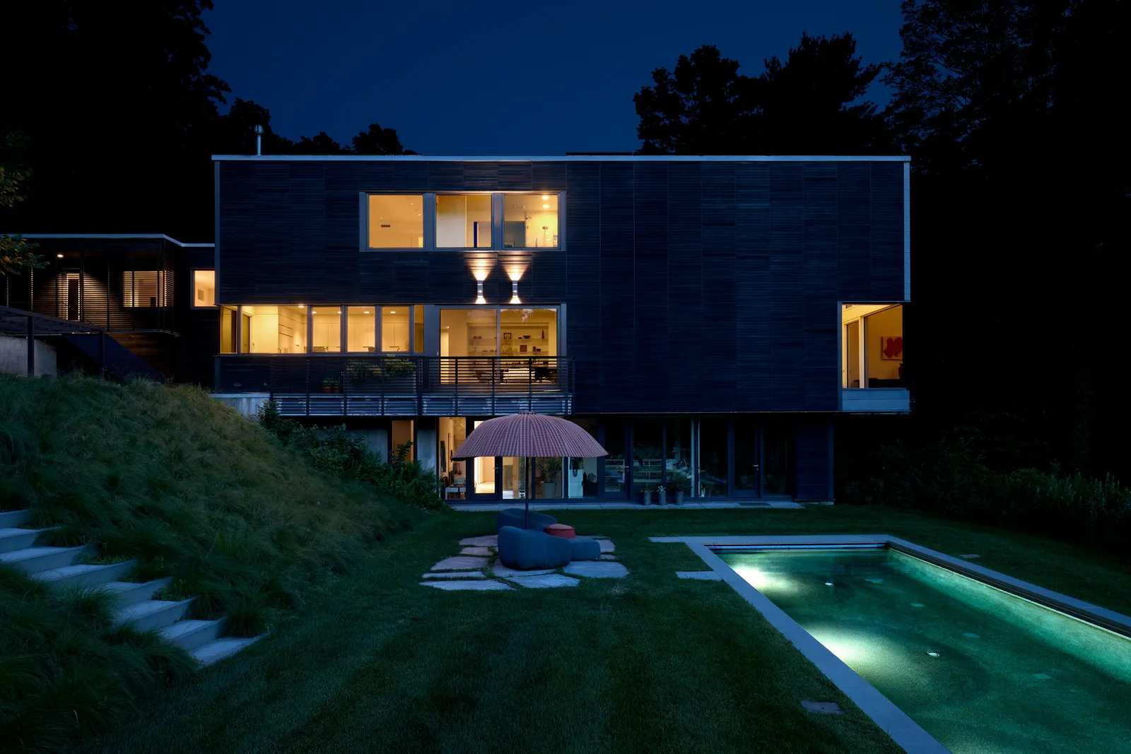 house at night with lit windows, a glowing pool, a pink umbrella and stone steps up the grassy slope