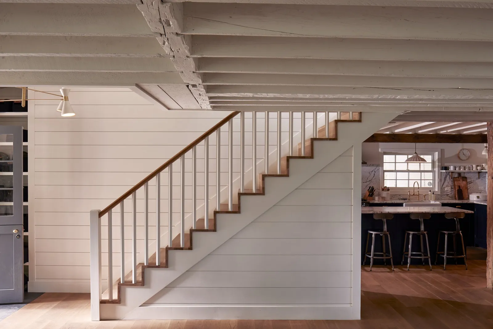 white-painted staircase with a wood handrail beside shiplap walls, the kitchen visible beyond